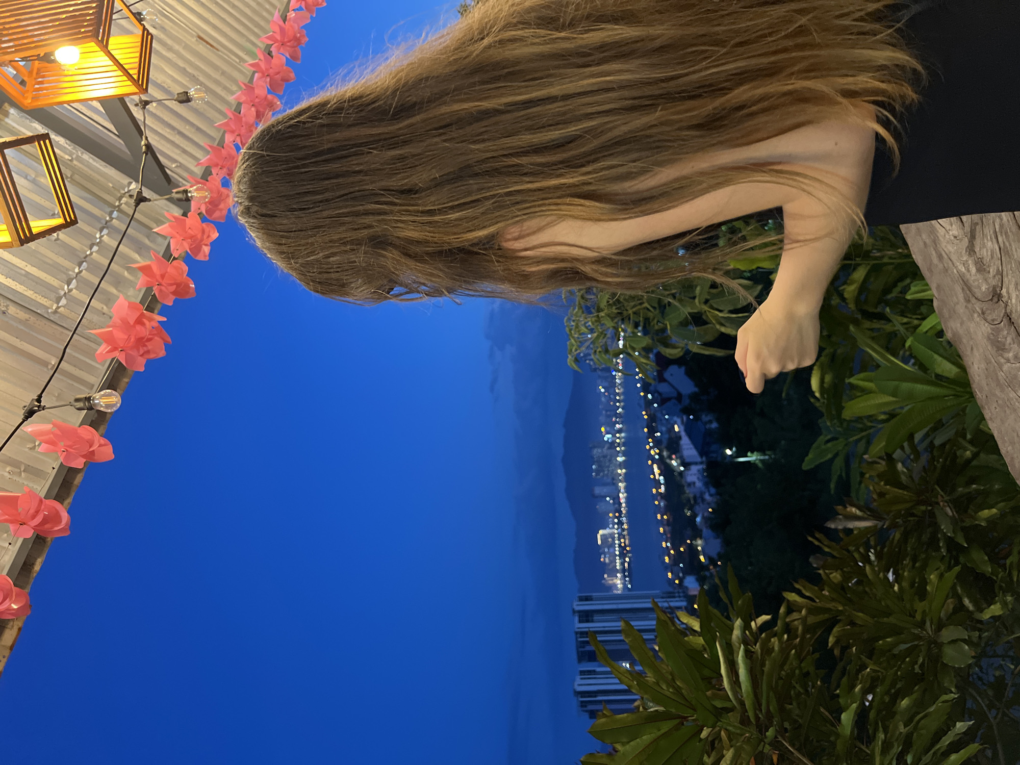 A medium shot captures the back of a young woman with long, light brown hair, wearing a black top, as she looks out at the cityscape of Nha Trang, Vietnam, at dusk. She stands on a balcony, with her right arm resting on a wooden railing. In the foreground, lush green foliage frames the view of the city, which is illuminated by numerous lights. The sky is a deep, vibrant blue, transitioning into darker hues as night approaches. Overhead, a string of pink paper flowers and decorative wooden light fixtures adorn the balcony's roof, adding a festive touch. The distant mountains form a silhouetted backdrop against the twilight sky. The overall scene conveys a sense of tranquility and awe as the city lights begin to twinkle.
