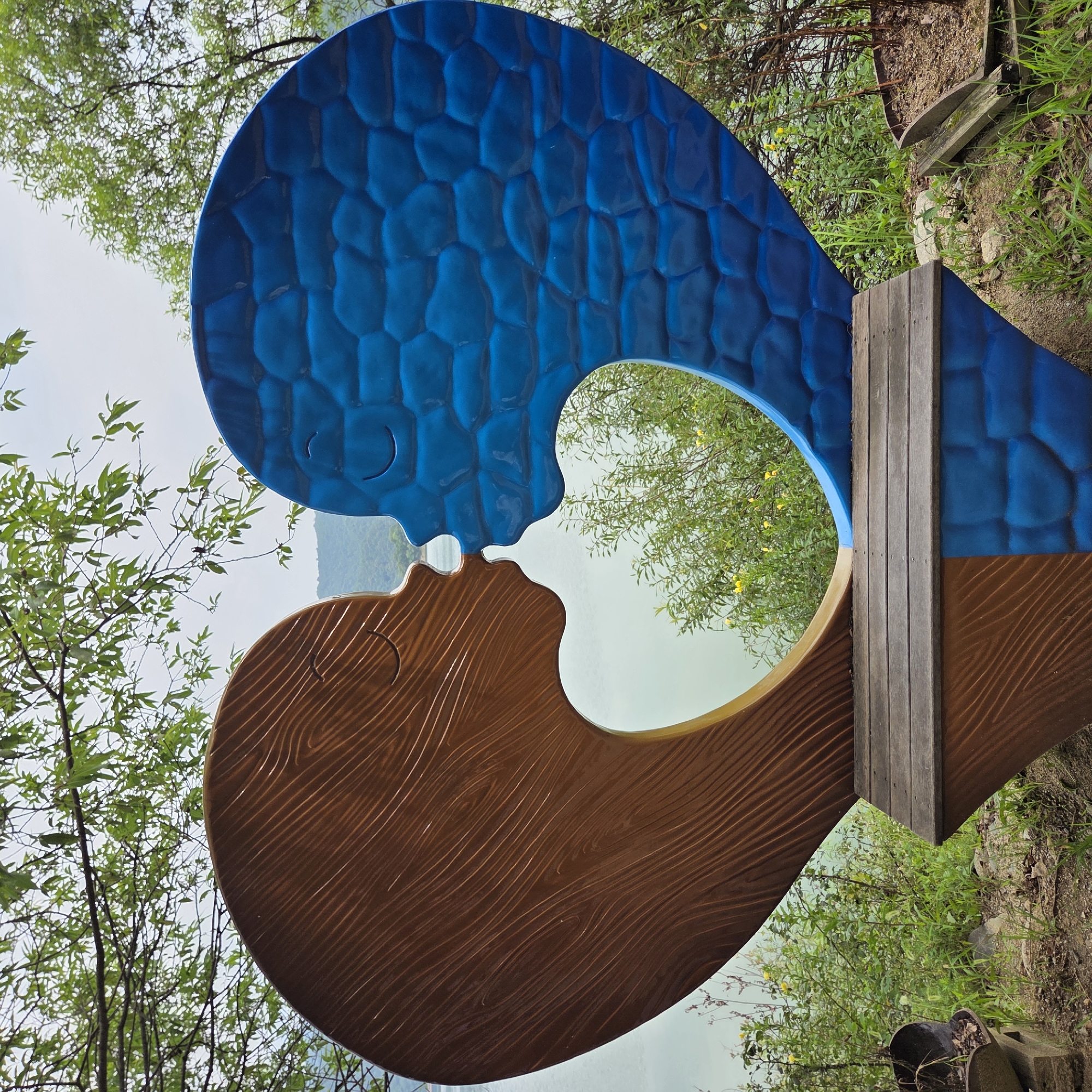 The image depicts a heart-shaped sculpture featuring two profiles facing each other in a kissing pose. The left profile is brown and textured to resemble wood grain, while the right profile is blue and has a scaled or faceted texture. Both profiles have closed eyes and smiling mouths. The sculpture is situated outdoors, with a body of water and distant hills visible in the background. Lush green foliage, including trees with visible branches and leaves, surrounds the sculpture. A wooden bench is integrated into the base of the sculpture. The ground in front of the bench is covered with dirt, grass, and some rocks. A broken planter with dirt and dead plant matter is visible to the left of the bench. The overall setting suggests a park or scenic outdoor location, likely in Gapcheon-myeon, South Korea, given the context provided. The lighting appears to be overcast, casting a soft, diffused light on the scene. There are no people present in the image.