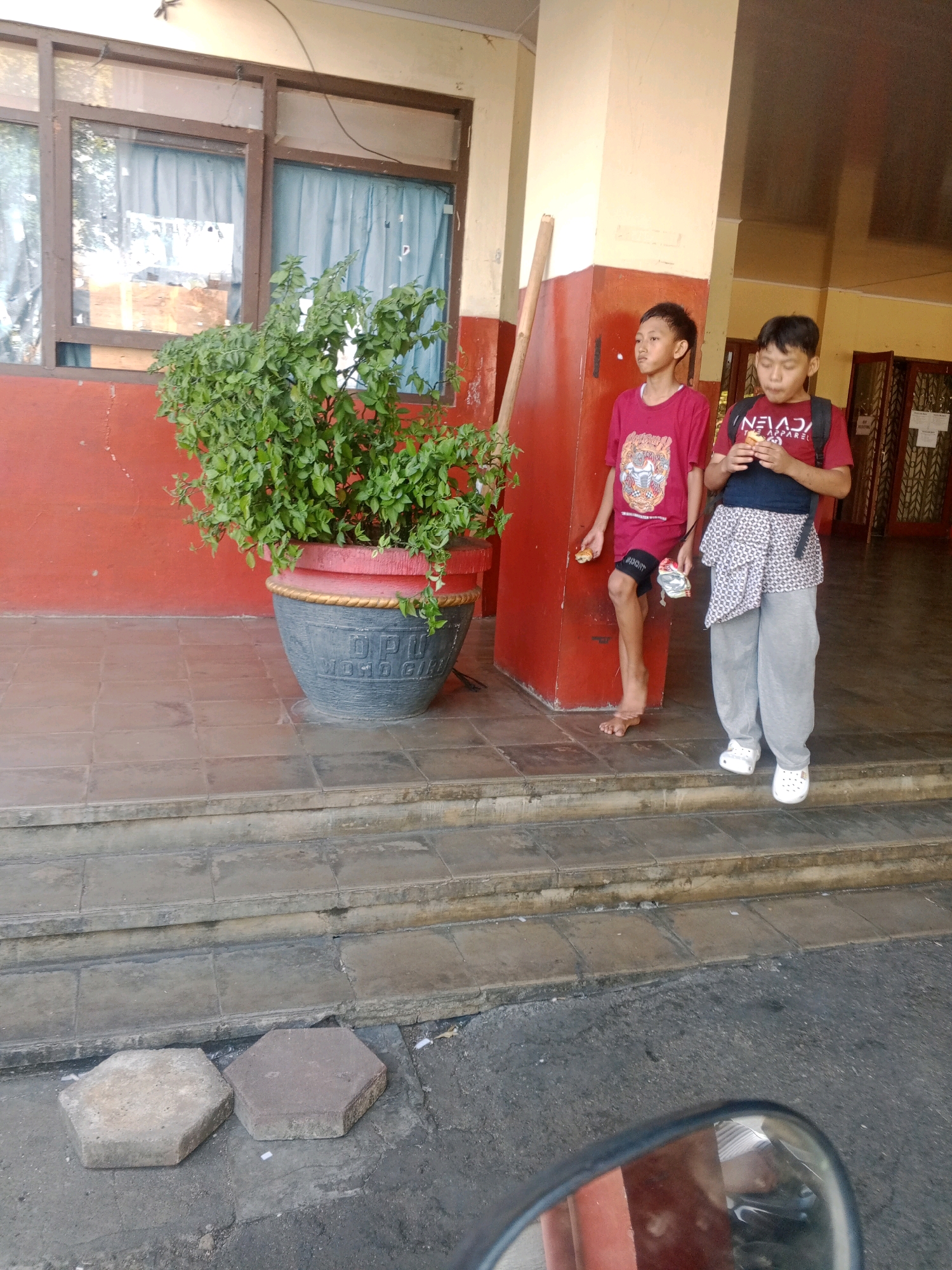 In Wuryorejo, Indonesia, two young boys stand on a tiled stairway outside a building with red walls and large windows. The boy on the left, wearing a red t-shirt and shorts, stands barefoot. The boy on the right, also in a red t-shirt and gray pants, wears white clogs and has a backpack. Both boys are holding small food items. A large potted plant with lush green leaves sits to their left. The stairway leads up from a paved area where two hexagonal concrete pavers lie on the ground. A portion of a motorcycle's rearview mirror is visible in the foreground.