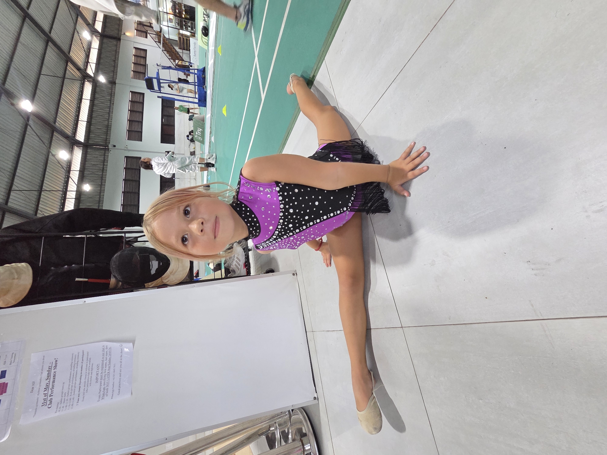 A young girl in a sparkly purple and black leotard, possibly a gymnastics or dance outfit, is performing a split on a grey tile floor. The environment suggests an indoor sports facility or a hall, as indicated by the green floor markings typical of a badminton or other similar court. In the background, there are fencing equipment and people in fencing attire. Also visible are stadium-style seating or bleachers. A whiteboard is positioned to the left, with printed announcements, including information about a "31st of May, Sunday - Club Performance Show!" and mentions WhatsApp number for registration indicating the city of North Kuta, Indonesia.