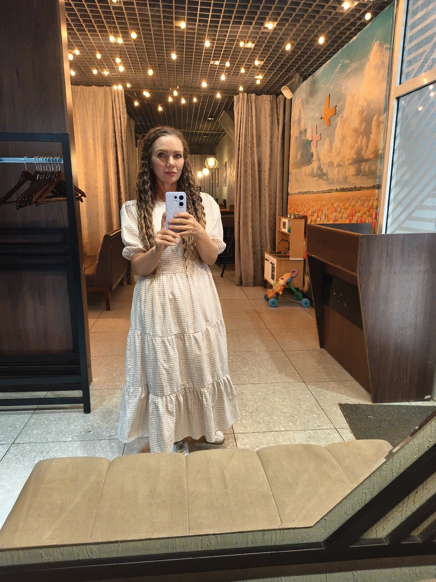 A woman with long, curly brown hair is standing and taking a selfie in a room with a mirrored wall and string lights on the ceiling. She is wearing a long, light-colored gingham dress with puffed sleeves. In the background, through the reflection, a rack of hangers is visible to the left, and a wall mural depicting a field of flowers under a cloudy sky is on the right. A child's toy kitchen and a small ride-on toy are also present in the background. To the right, a dark wooden counter or desk is visible. In the foreground, a padded bench is partially visible. The floor is tiled.