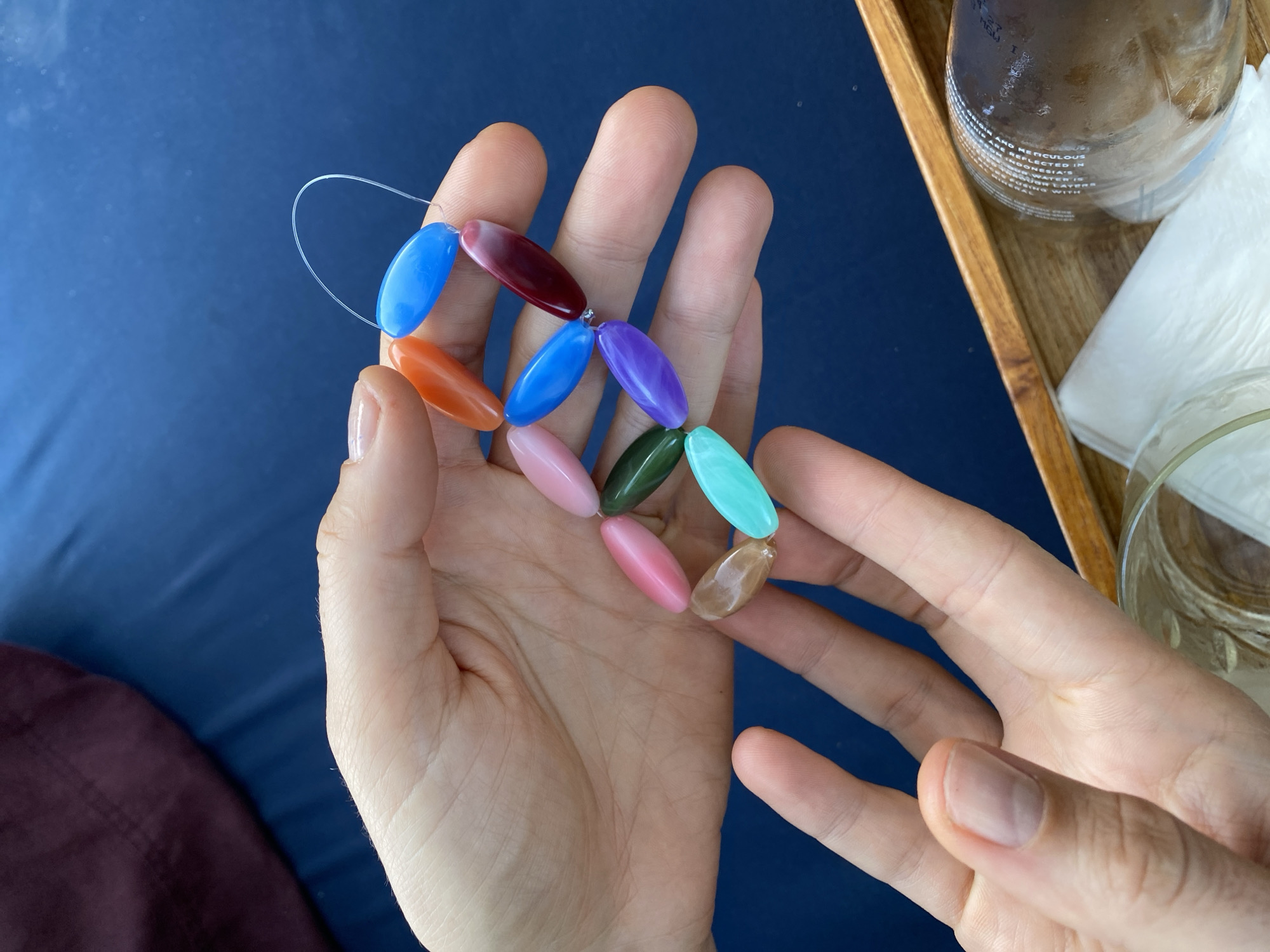 Two hands are holding a strand of oval-shaped beads of various colors: orange, blue, pink, dark red, green, purple, and turquoise. The beads are strung together with clear thread, forming a loose curve. The hands are pale-skinned with short fingernails, one hand displaying a light pink nail polish on the thumbnail. The background consists of a dark blue surface, possibly a bed or sofa, with a section of dark burgundy fabric to the upper left. A wooden tray is visible in the lower right corner, containing a clear glass and a frosted glass container with text. The image appears to have been taken indoors.