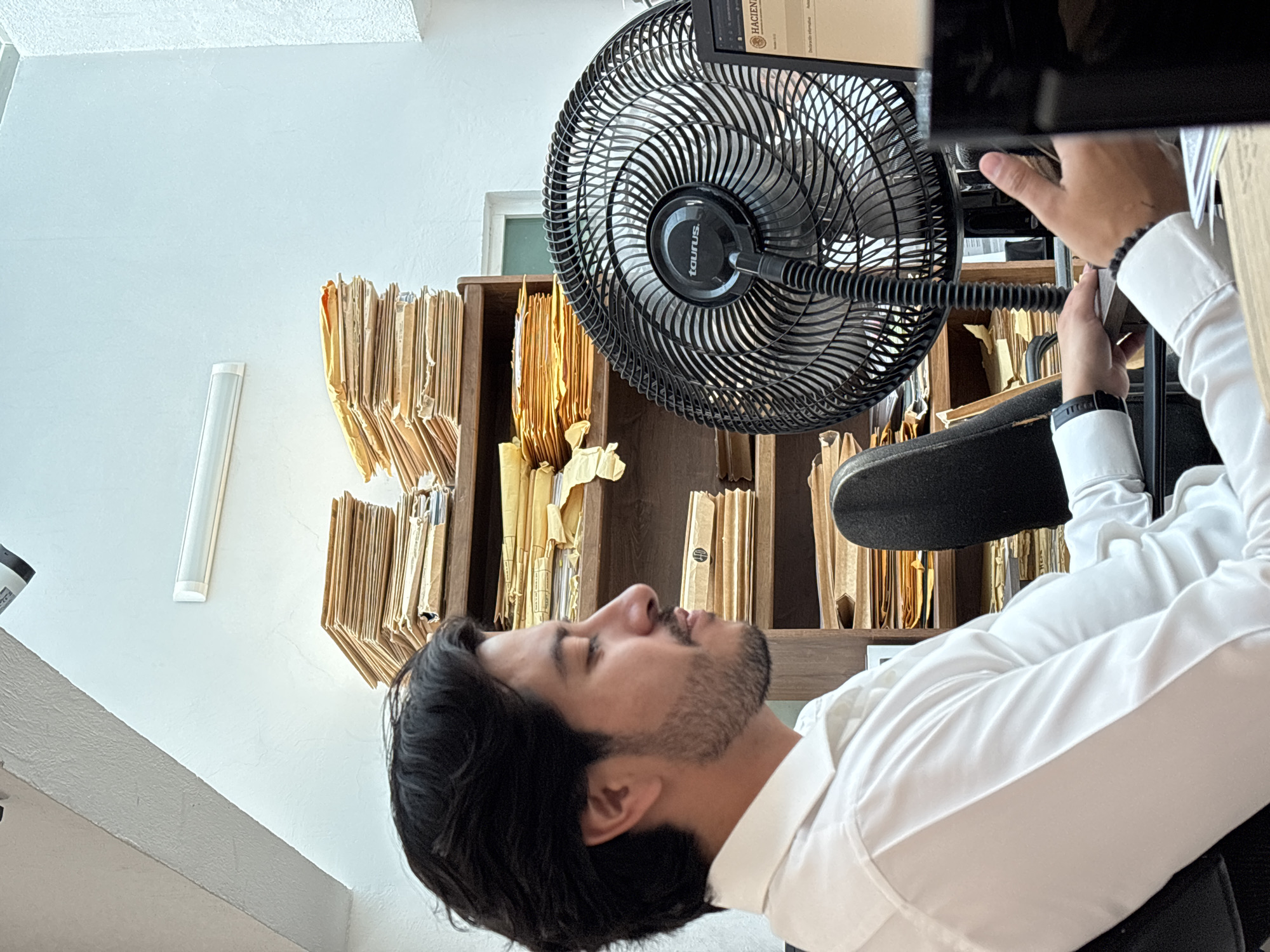 A person in a white shirt is seen from the side. Their head is tilted back towards an office fan. To the left and behind the person is a shelf filled with golden brown paper folders. The image was taken indoors in an office environment. The location is Puebla, Mexico.