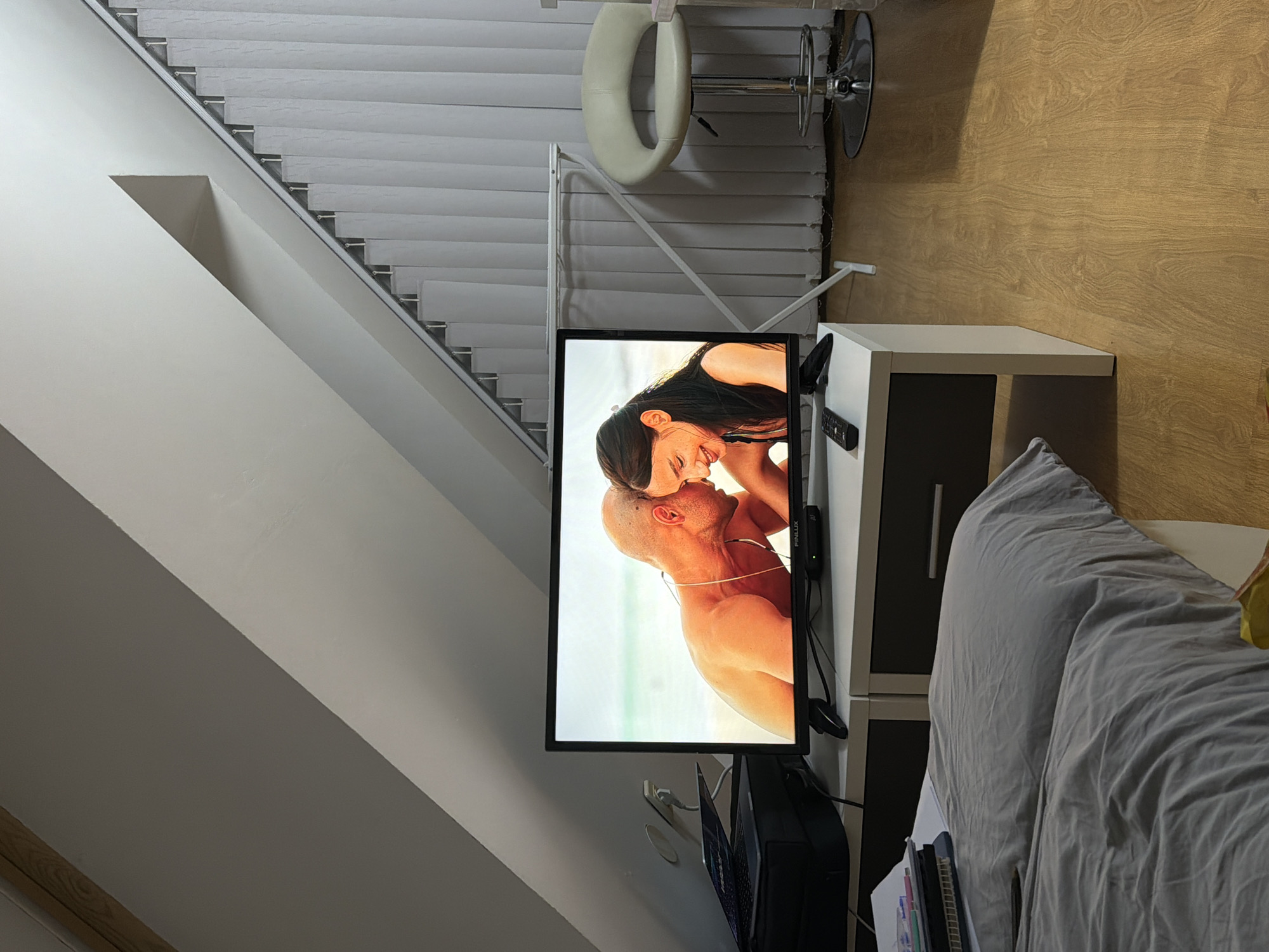The image shows an interior room setting. A television is displaying a romantic scene of a couple embracing against a background of what appears to be a beach. To the right of the TV is a white and dark grey TV stand. A grey comforter is partially visible to the right of the TV stand. In the background there are white window blinds and a white barstool is nearby. The floor is wood. The location is not possible to determine with the available visual information, but it may be possible to confirm that with another image of the exterior and interior.