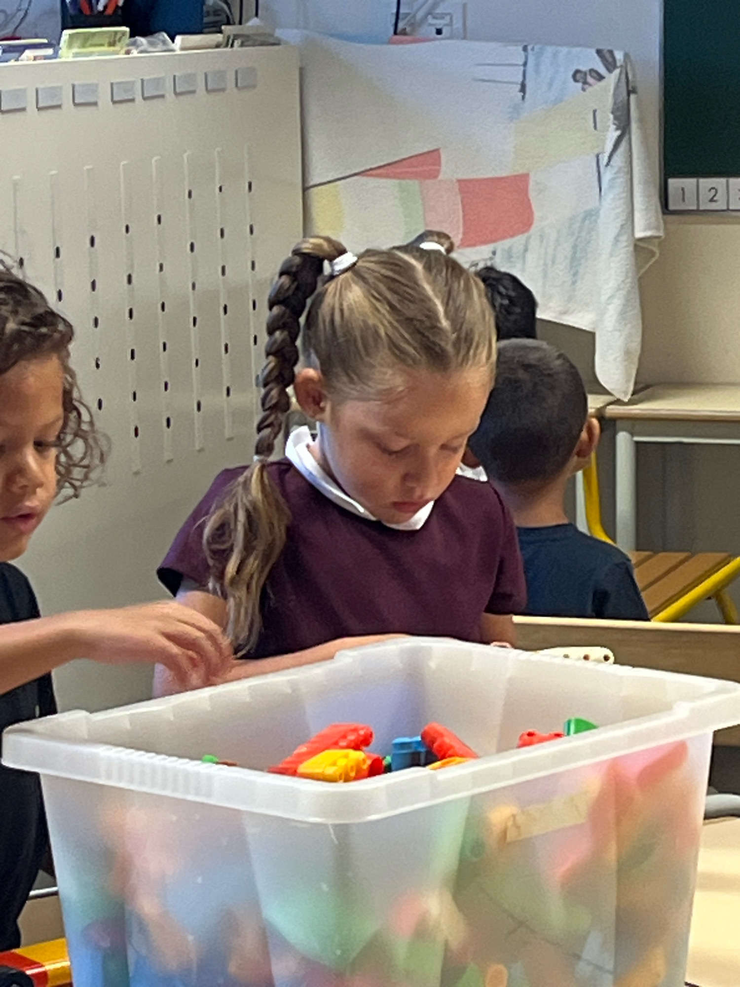 The image shows a classroom in La Fouillouse, France. In the foreground, a young girl with blonde hair in a braid is looking down at a clear plastic bin filled with colorful toys. To her left, another child with curly dark hair is partially visible, reaching towards the bin. Behind the girl, a young boy with dark hair is facing away from the camera. The background includes classroom elements such as shelves, a bulletin board with fabric decorations, and a green board with numbers. The lighting suggests an indoor setting, likely during daytime.
