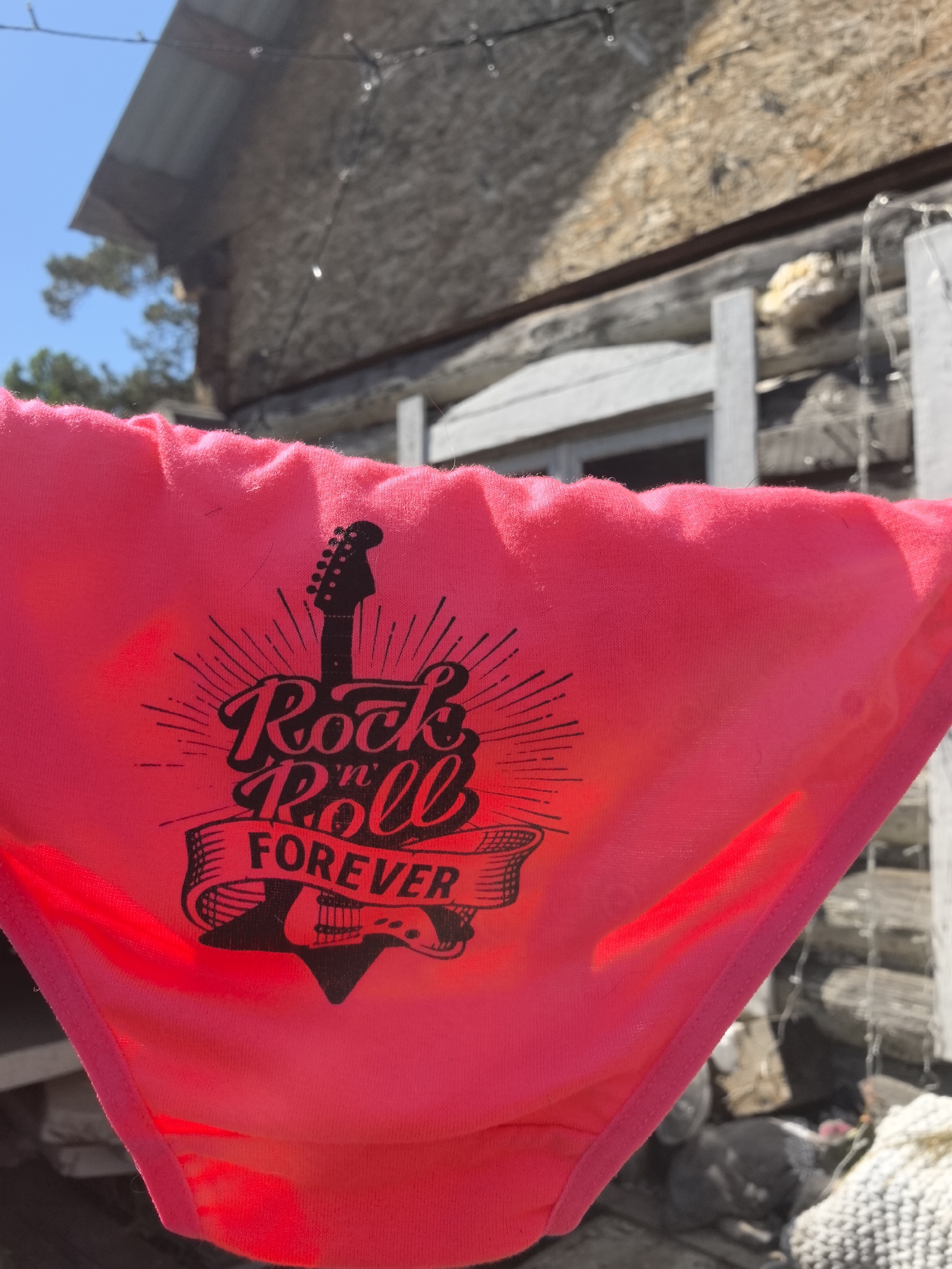 A pink garment with a "Rock 'n Roll Forever" design is prominently displayed. The design features a guitar silhouette and stylized lettering. In the background, an out-of-focus rustic wooden building is visible under a bright blue sky. The precise location is not discernible based on the image alone. The image does not provide sufficient details to determine if the location is in the city of Noviy, Russia.