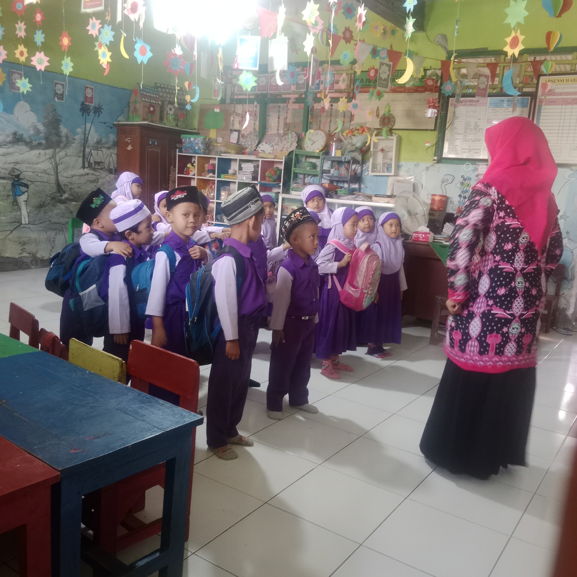 In Tegowanu Kulon, Indonesia, a classroom scene unfolds with a group of young children dressed in purple uniforms and headscarves, some wearing black peci hats. The children are standing in front of a woman wearing a bright pink hijab and a patterned black and pink top, facing away from the camera. They appear to be a class, possibly preparing for an activity or lesson. The classroom walls are decorated with colorful paper cutouts of stars, moons, and suns, along with educational charts and artwork. Shelving units filled with books, toys, and other classroom supplies are visible in the background. The floor is tiled with white square tiles. The overall impression is of a vibrant and educational environment for young learners.