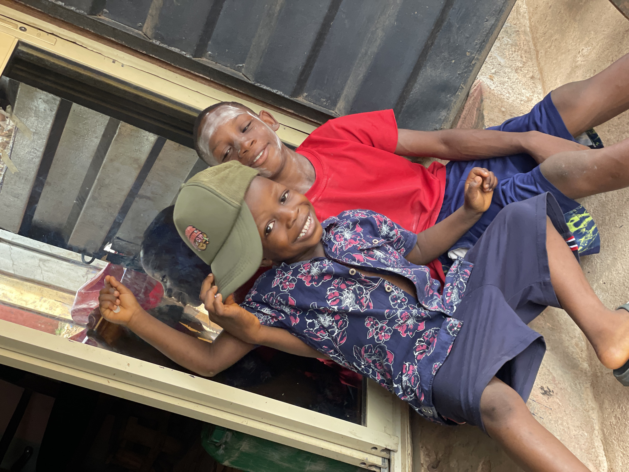 Two young boys are lying down side-by-side in what appears to be a window frame or ledge. One boy wears a red t-shirt and blue shorts, and has a white mark on his forehead. The other boy wears a floral-patterned navy shirt, blue shorts and a green baseball cap. Both are smiling. The background includes a dark metal or painted surface. The location is presumed to be in the city of Onitsha, Nigeria.