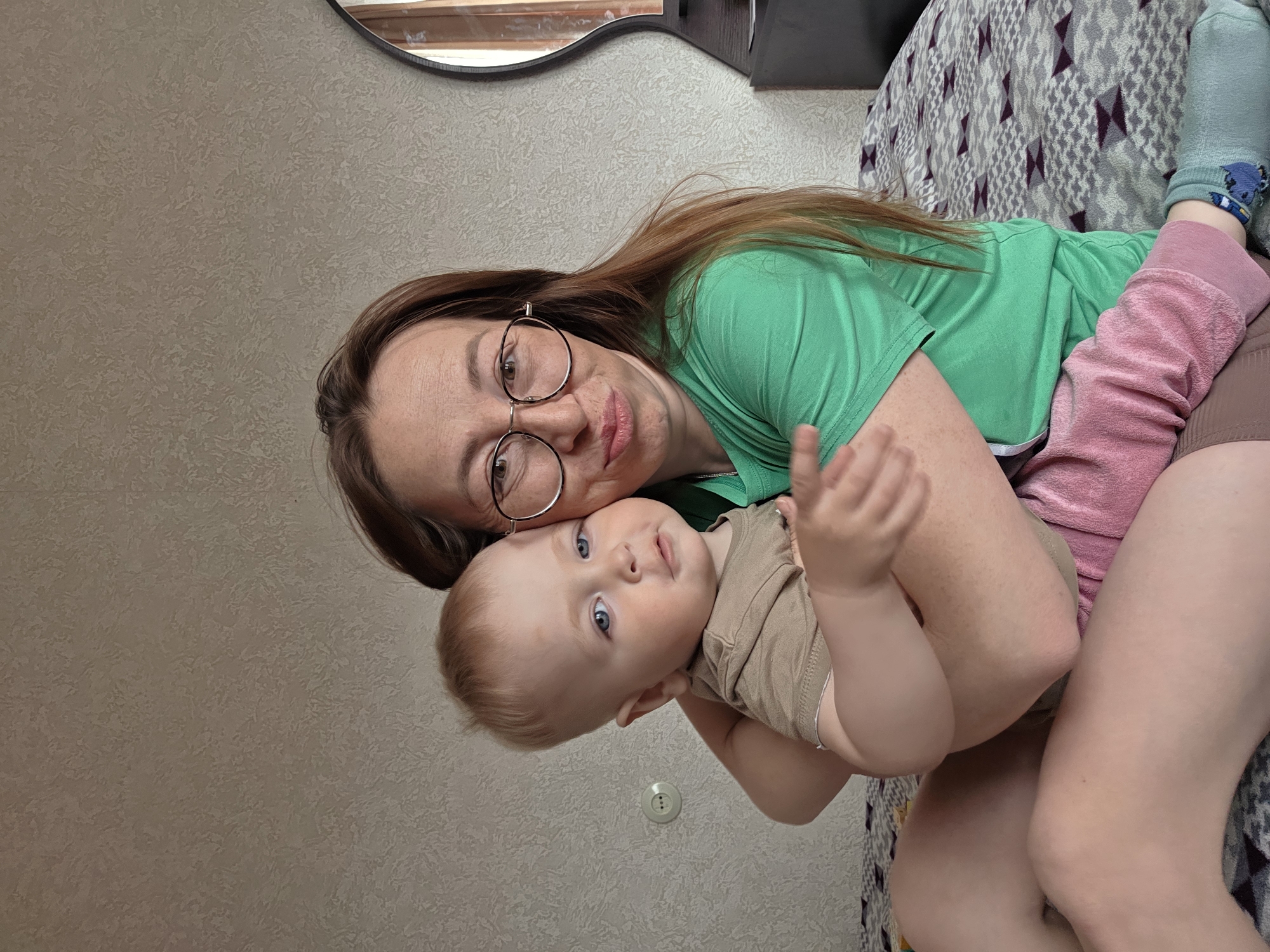 A woman with auburn hair, wearing glasses and a green t-shirt, is lying on a bed, holding a baby in her arms. The baby is wearing a brown t-shirt and pink bottoms, with a blue sock on one foot. The bed is covered with a patterned blanket. A circular mirror is visible on the wall behind the woman. The setting appears to be indoors, potentially in a home. The image was taken in Chaikovsky, Russia.