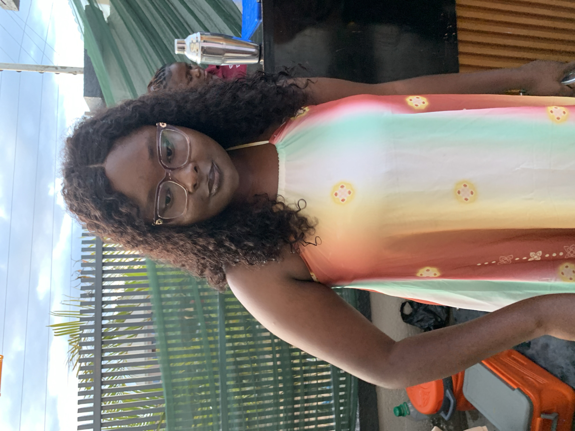A medium shot captures a woman with curly dark hair and glasses posing outdoors in Gura Top, Nigeria. She is wearing a sleeveless, tie-dye dress with a pastel gradient of yellow, white, green, and red, adorned with small yellow circular floral patterns. She has dark lipstick and her gaze is directed downwards. To her right, a person in a maroon top and another individual are partially visible behind a stainless steel cocktail shaker and a blue tray on a counter. In the background, a green metal fence and palm fronds are visible, suggesting an outdoor establishment. To the left of the woman, two orange cooler boxes and a plastic water bottle with a green cap are on the ground. The sky above is overcast.