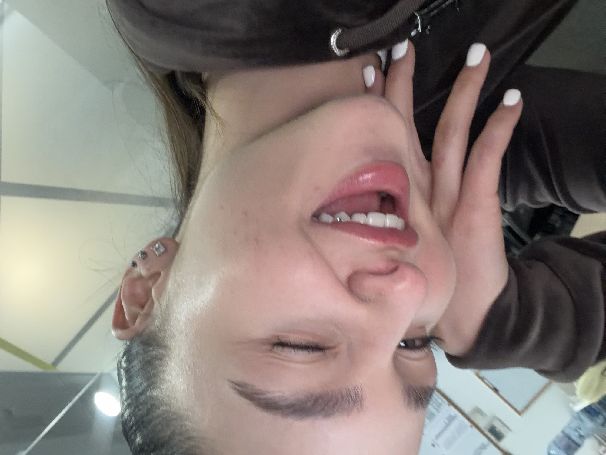 A close-up, upside-down image shows a young woman with fair skin, brown hair, and manicured fingernails with white polish. She is wearing a brown velvet hoodie. Her mouth is open, showing teeth. She has piercings in her ear. The background contains a white ceiling and a partially visible wall with papers attached. The location is indeterminate; there is no visible indication of the city of Sofia, Bulgaria.