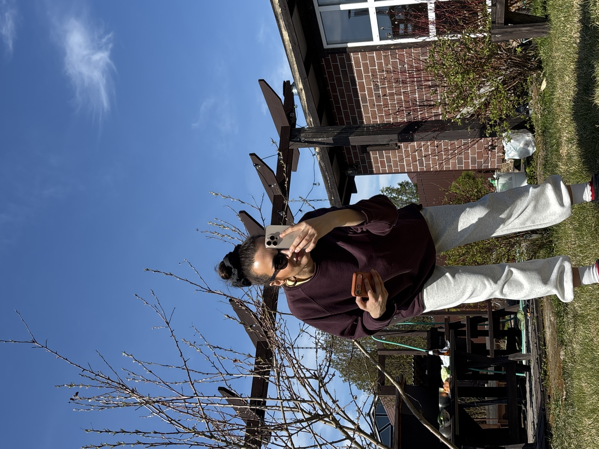A woman is standing outdoors in an area with a lawn and a wooden structure, possibly a pergola. She is holding a smartphone in one hand, apparently taking a picture, and a smaller object in the other hand. She is wearing a dark-colored sweatshirt and light-colored sweatpants. A tree with bare branches is visible in the foreground. In the background, there's a brick building and a blue sky with wispy clouds. The location is likely in Emel'yanovo, Russia, given the request.