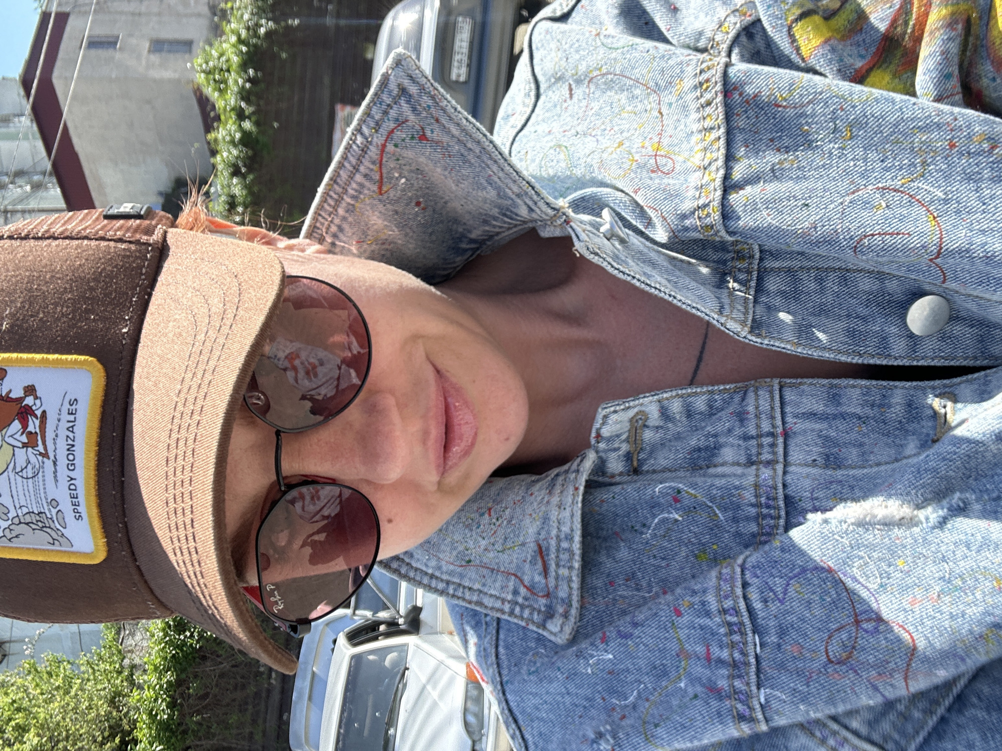 A person wearing a brown baseball cap with a "Speedy Gonzales" patch, sunglasses, and a light blue denim jacket is pictured in an outdoor setting. The person is positioned at an angle towards the camera. Part of a white vehicle is visible in the background, as well as some greenery and a building. The location is unspecified.