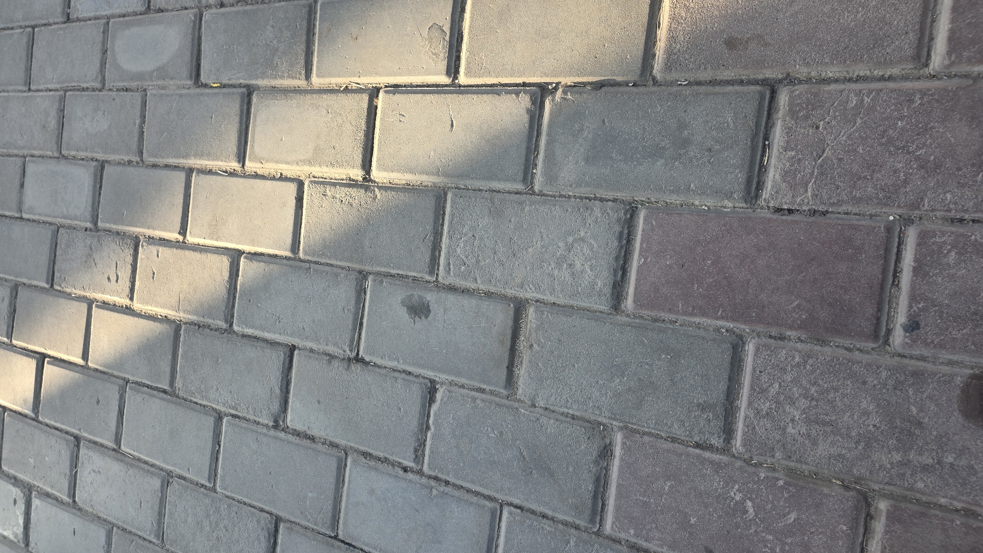 The image depicts a paved surface composed of rectangular interlocking bricks, arranged in a staggered pattern. The bricks vary in color, with some appearing grey and others a reddish-brown hue. Sunlight casts diagonal shadows across the pavement, indicating it is outdoors. The ground shows signs of wear and tear, with some dirt and debris accumulated between the bricks. The scene is likely a walkway or pedestrian area in Nariman, Kyrgyzstan, given the context. There are no people or specific objects of interest visible in the frame, beyond the paving stones themselves. The focus is entirely on the texture and pattern of the ground.