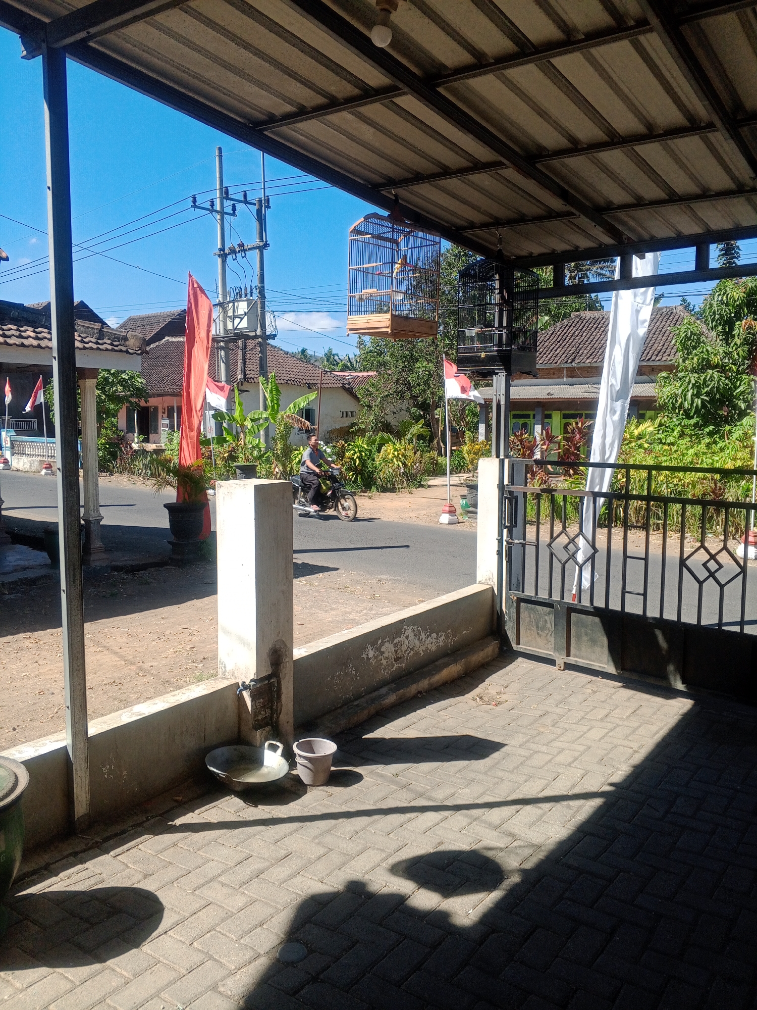 The image depicts a view of a street in Kemulan, Indonesia, looking towards residential buildings under a clear blue sky. In the foreground, a covered outdoor area with a corrugated metal roof is visible, supported by metal beams. Two birdcages are hanging from the ceiling, one containing a bird. A black metal gate is in the right foreground.

On the street, a man is riding a motorcycle. Red and white flags, likely the Indonesian flag, are planted on flagpoles outside a house. Various potted plants are scattered around the area. A transformer and utility poles with electrical wires are present on the side of the road.

In the immediate foreground, a low concrete wall runs horizontally, with a metal pipe and faucet attached to a concrete pillar. A metal basin filled with water and a small plastic bucket are placed on the paved ground next to the wall. The ground is paved with interlocking bricks, and shadows from the overhead structure and objects are cast upon it. The scene appears to be a residential street in a tropical climate.