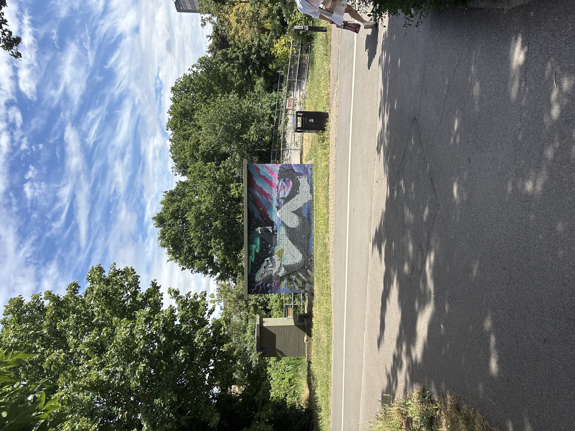 A person is walking on a paved path next to a grassy area with trees and a mural. The mural depicts a stylized creature with wings and a human-like face against a background of colorful stripes and geometric patterns. The setting appears to be outdoors, possibly a park or urban green space, given the presence of trees, a paved path, and a trash can. The time of day is likely daytime, as indicated by the bright lighting and the shadows cast by the trees. The weather seems to be fair, with a blue sky and wispy clouds visible. The person is wearing white shorts and a white top, and appears to be carrying a bag. The overall mood is relaxed and casual. There is no visible text in the image.