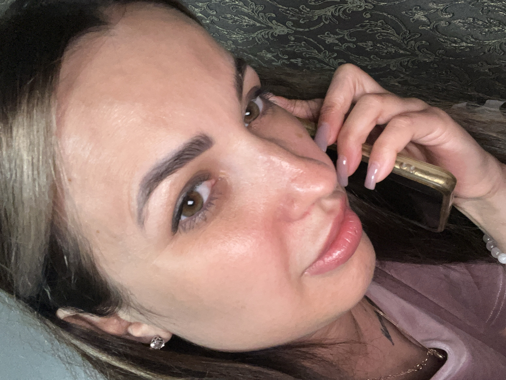 A close-up, over-the-shoulder selfie shows a woman with dark hair, light skin, and brown eyes holding a smartphone to her ear. She is wearing a pink shirt and a delicate necklace. Her fingernails are painted a light lavender color. The background is dark and textured, with a subtle floral pattern. The lighting is soft and appears to be artificial. The woman's expression is neutral, and she is looking slightly away from the camera. The image was taken in an indoor environment.
