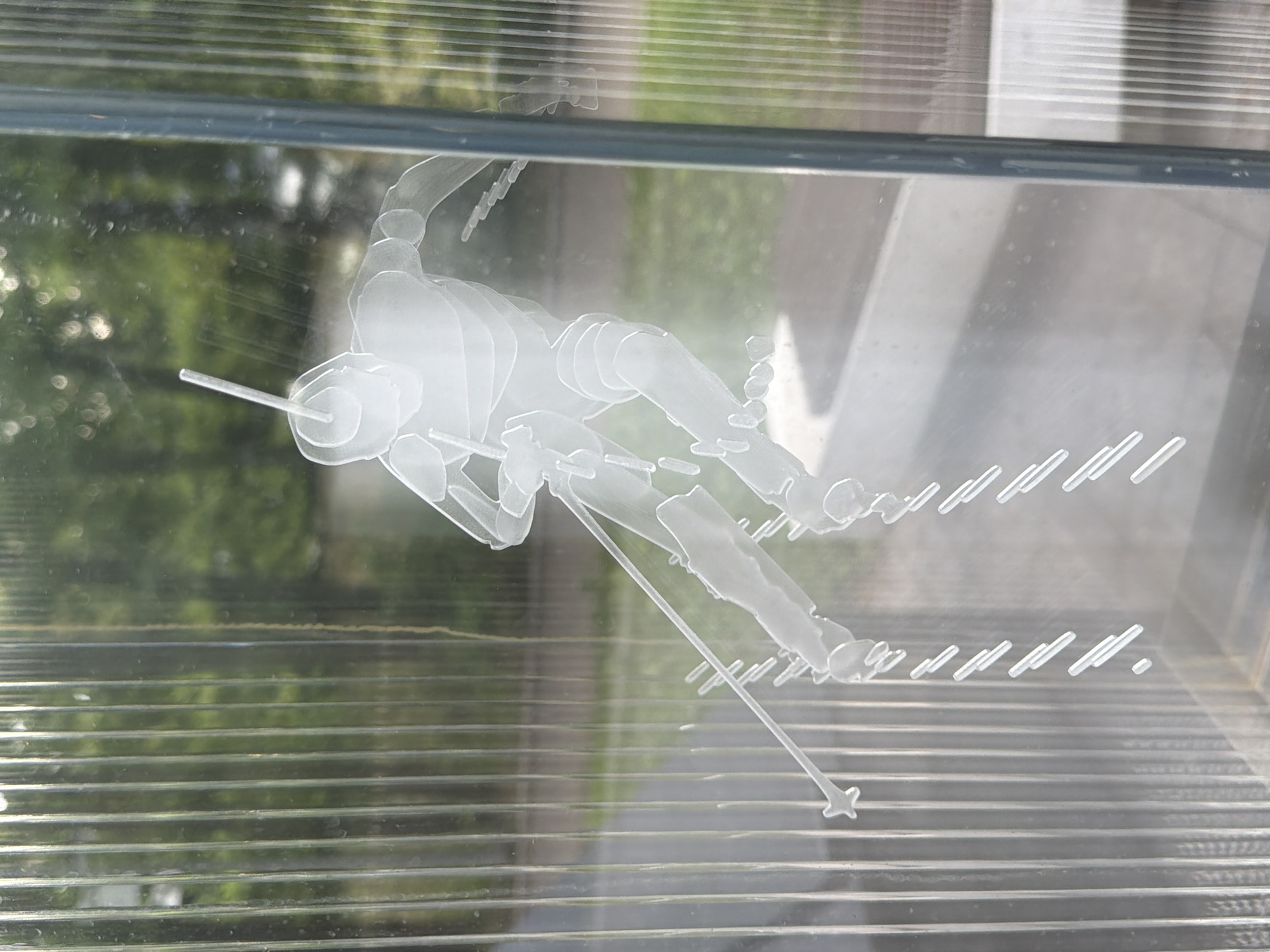 An etched glass panel depicts a silhouette of a skier in motion. The skier is positioned in a dynamic pose, with skis extending forward and poles held out. The etching is white against a clear background, with vertical ribbed lines of the glass panel forming a backdrop. The scene appears to be outdoors, with blurred greenery and hints of light visible through the glass, suggesting trees or foliage. The glass panel itself is framed and positioned in an upright manner, likely part of a larger structure or exhibit. The location is unknown.