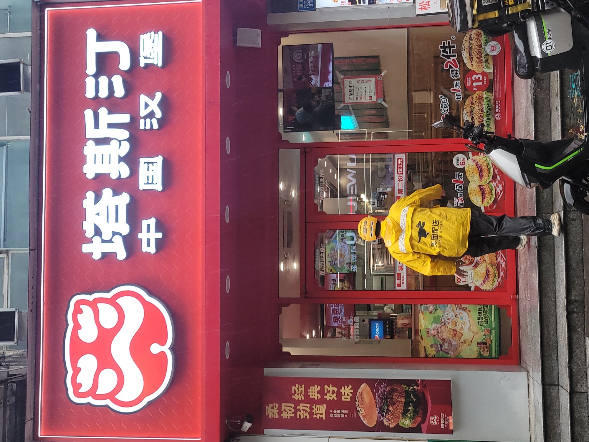 The image captures a vibrant street scene in front of a "Tasiting Chinese Burger" (塔斯汀 中国汉堡) fast-food restaurant in Liangjiang, China. The restaurant features a prominent red facade with white, illuminated Chinese characters spelling out its name, and a distinctive red and white logo resembling an abstract animal paw print.
A delivery person, wearing a bright yellow uniform with the "美团配送" (Meituan Delivery) logo and a bird emblem, stands directly in front of the glass entrance doors, likely waiting for or having just completed an order. A black and white scooter, possibly belonging to the delivery person, is parked nearby.
The large glass windows and doors of the restaurant are adorned with various advertisements and promotional posters. Visible text includes offers like "加1元得2件" (Add 1 yuan for 2 items) and "第二份仅1元" (Second item only 1 yuan), alongside images of appetizing burgers, some priced at "¥13". Other signs mention "会员日" (Membership Day), "广纳贤才" (Recruiting talents), and a promotion featuring "萌兽组团 山野寻堡" (Cute beasts group up, mountain wilderness burger hunt). A Pepsi logo is also visible inside. On the left side, posters advertise "柔韧劲道 经典好味" (Tender and chewy, classic good taste) and "香辣鸡腿堡" (Spicy chicken leg burger).
The scene appears to be daytime, possibly overcast or late afternoon, as the neon signs are lit. Subtle streaks on the glass suggest recent or ongoing light rain. The overall atmosphere is that of a bustling urban environment with ongoing commercial activity. Additional small signs on the adjacent wall show "19号" (No. 19), "松" (Song), and "招" (Recruitment).
