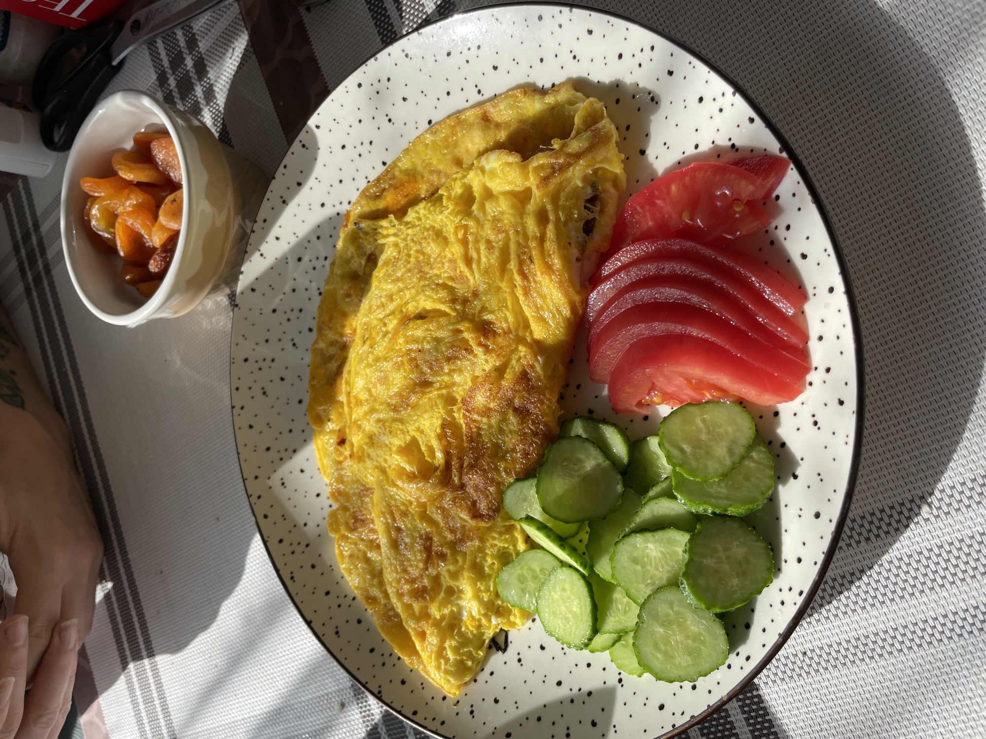An overhead, eye-level shot captures a breakfast spread laid out on a white and grey checkered tablecloth. The primary focus is a large, golden-yellow omelet resting on a speckled white plate with a dark rim. To the left of the omelet are thinly sliced, vibrant green cucumbers, and to its right, several fanned-out slices of bright red tomato. In the upper right corner of the frame, a small, light-colored bowl contains a pile of dried apricots. In the background, partially out of focus, are a pair of human hands, appearing to be of an adult, with visible fingernails and a tattoo on the wrist of the left hand. A dark-handled pair of scissors is also visible in the upper right quadrant. The scene is bathed in natural light, casting distinct shadows across the tablecloth. The image does not provide any specific information about the location in the city of Kornevo, Russia.