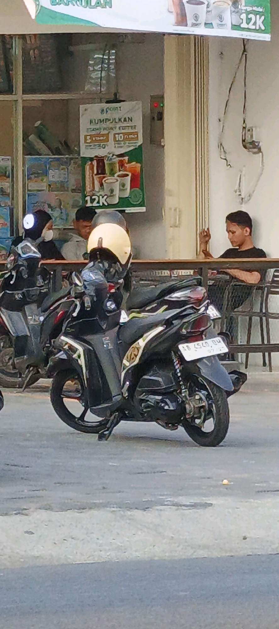 In Sidoarum, Indonesia, a street scene unfolds with several individuals and motorcycles. In the foreground, a black motorcycle with yellow accents and a light-colored helmet is parked. Behind it, another black motorcycle is partially visible. Three men are seated at a table in what appears to be an outdoor seating area of a cafe or shop. One man, wearing a black t-shirt, is on the right, looking downwards with his arm resting on the table. To his left, another man in a grey hoodie is facing forward, and a third man, partially obscured by the motorcycle and wearing a dark headscarf and a white mask, is also seated. A banner hangs in the background with Indonesian text and images of drinks, indicating a promotion or advertisement. The environment suggests a public space with paved ground and architectural elements typical of Indonesian street scenes.