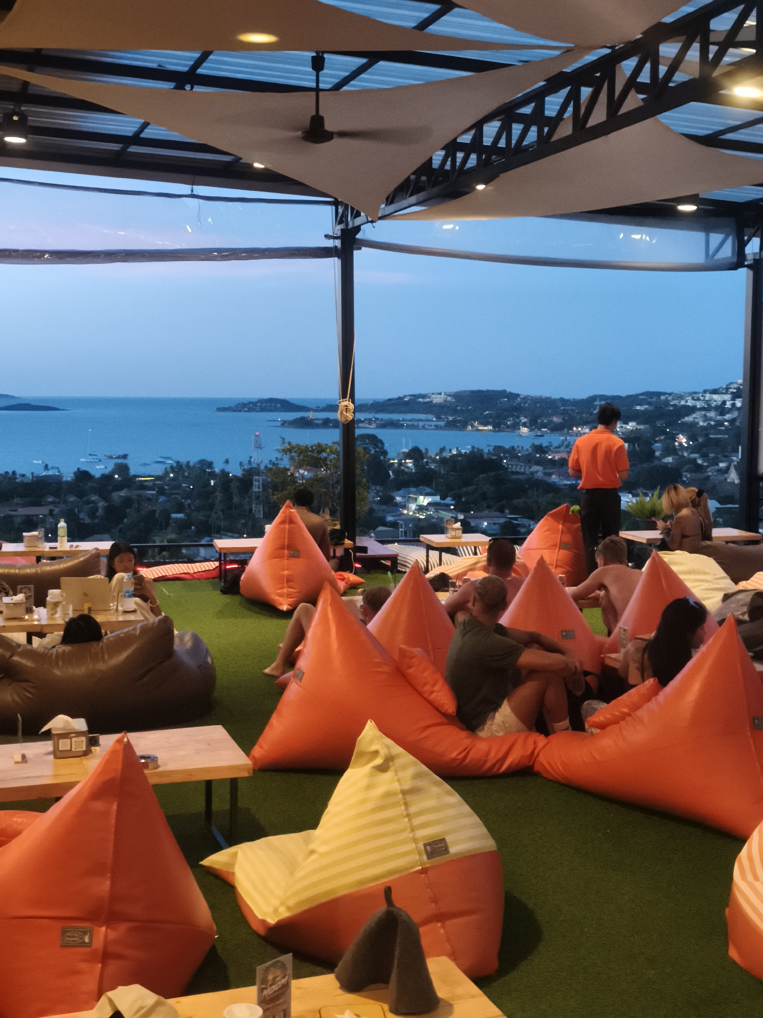 The image depicts a rooftop bar or restaurant at dusk in Ban Rak, Thailand, with a panoramic view of the ocean and surrounding hills. The foreground is filled with numerous large, colorful beanbag chairs arranged on a green, artificial turf surface. People are seated and lounging on these beanbags, some interacting, others looking out at the view. A few low tables are scattered among the seating. In the middle ground, a man in an orange shirt stands looking out at the scenery. Further back, the ocean stretches to the horizon, dotted with boats, and the coastline is lined with buildings and lush vegetation. The sky is a gradient of blue and orange, indicating the transition from day to night. A roof structure with fabric panels and lighting is visible overhead.