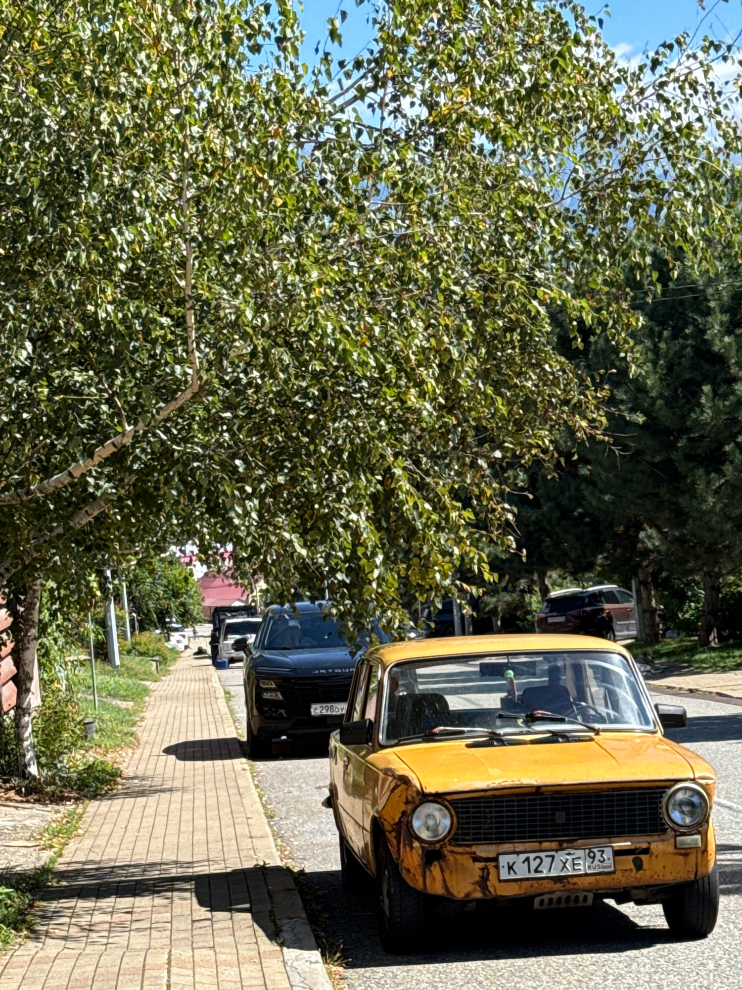 In Krasnaya Polyana, Russia, a weathered yellow car is parked on a paved road. The car has a license plate that reads "K 127 XE 93 RUS". Adjacent to the road is a brick sidewalk, lined with green foliage and trees. A black SUV is parked on the other side of the sidewalk. Further down the road, other cars and buildings are visible, suggesting a residential or commercial area. The sky above is blue with some scattered clouds, and the sun is casting shadows.