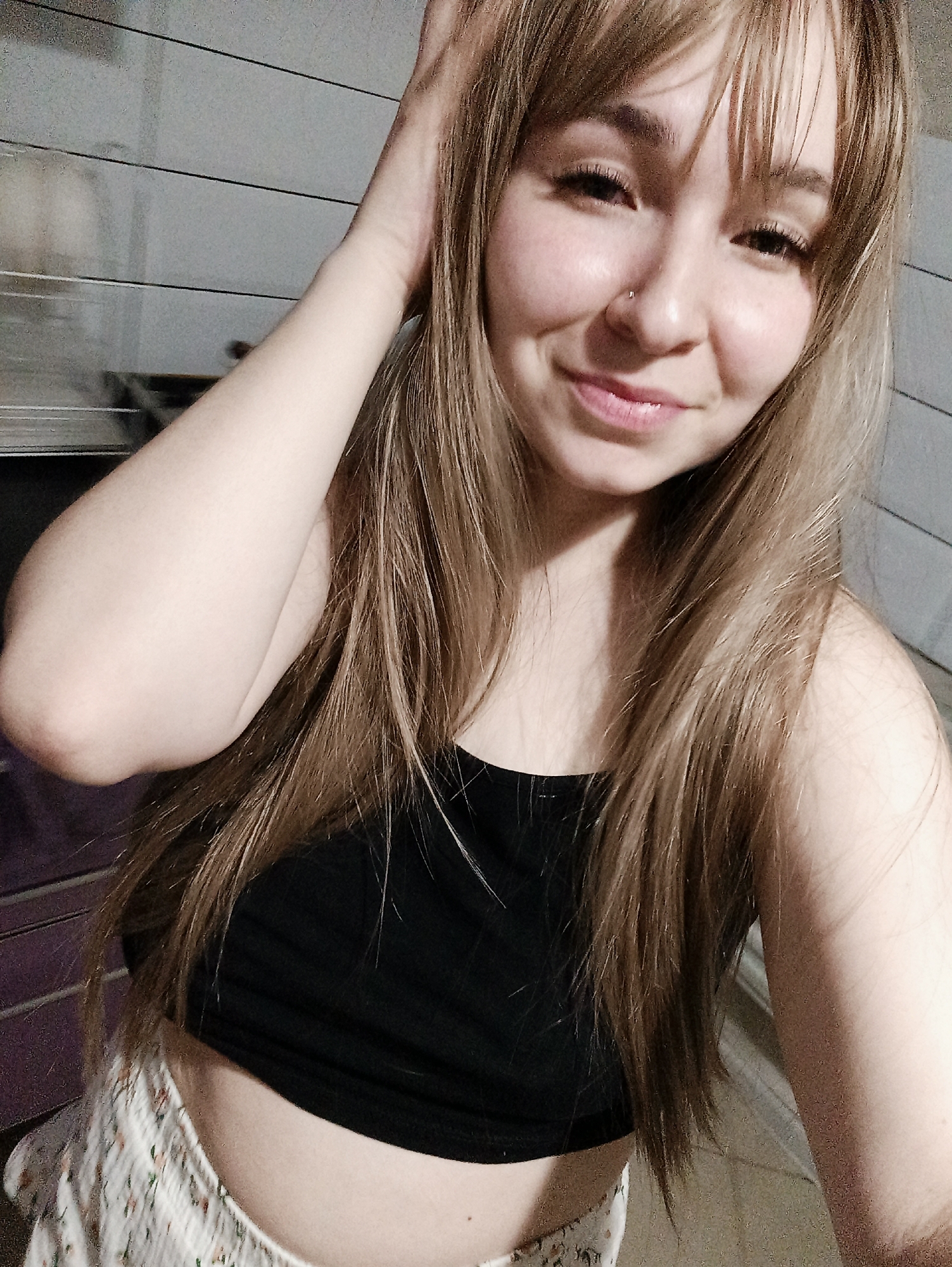 The image displays a close-up, eye-level portrait of a young adult female, identified as being located indoors in Izhevsk, Russia. The subject has fair skin, long light-brown hair with bangs, and a small stud piercing in her left nostril. She is depicted smiling directly at the viewer. Her right hand is raised, with fingers splayed, resting near the top of her head. She is wearing a black, strapless or spaghetti-strap crop top, revealing her midriff, and light-colored shorts with a subtle floral or patterned print. The background consists of a light-grey wall with distinct horizontal paneling or grooves. A darker, vertical surface, possibly purple, is visible on the far left side of the frame. The scene is illuminated by soft, frontal lighting.