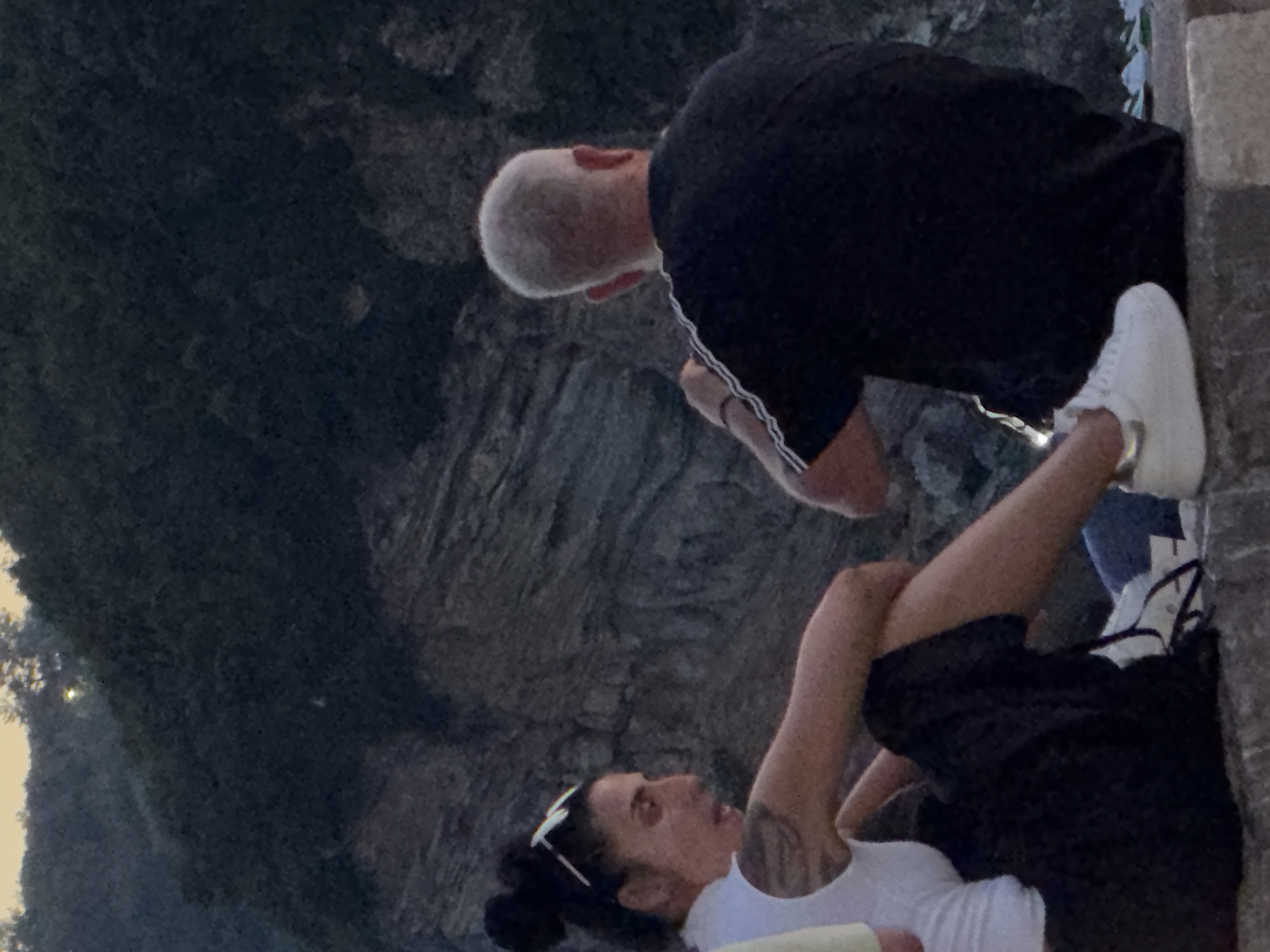 Two individuals, a woman and a man, are seated on a stone ledge, facing away from the camera in a medium shot. The woman, on the left, has her dark hair pulled back in a bun and wears white-rimmed sunglasses on her head. She is dressed in a white top and a dark, flowing skirt. A tattoo is visible on her left arm. The man, on the right, has short, light-colored hair and wears a dark t-shirt with white stripes on the sleeves. They appear to be in a casual setting, possibly outdoors. The background consists of a rocky, textured surface and foliage, suggestive of a natural landscape. The lighting indicates it is either dawn or dusk. This scene is set in Budva, Montenegro.