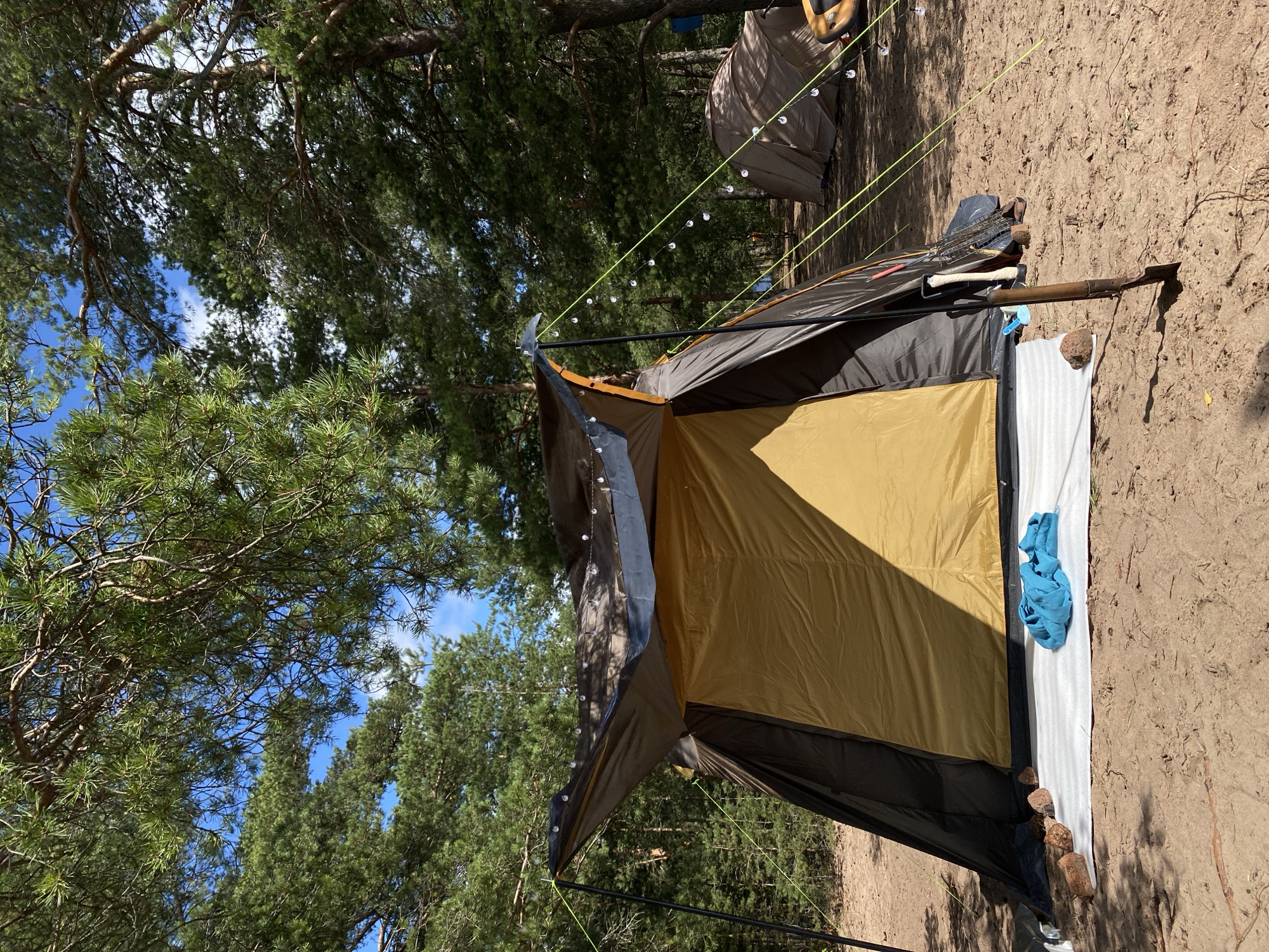 This image depicts a campsite in the city of Baltiiskoe, Russia, surrounded by a dense pine forest. A large, dark brown and gold tent is pitched prominently in the foreground. Its entrance is open, revealing the golden interior. A white ground tarp is spread in front of the tent, with a crumpled blue t-shirt lying on it. Several reddish-brown rocks are placed along the edge of the tarp. To the right of the tent, a shovel is stuck into the sandy ground. In the background, another beige tent is visible, partially obscured by trees, suggesting multiple campers are present. String lights with small bulbs are draped over the front of the main tent, extending towards the trees. The ground is sandy and littered with pine needles and small branches. The sky above is blue with scattered white clouds, and the sun casts shadows from the trees and tent poles.