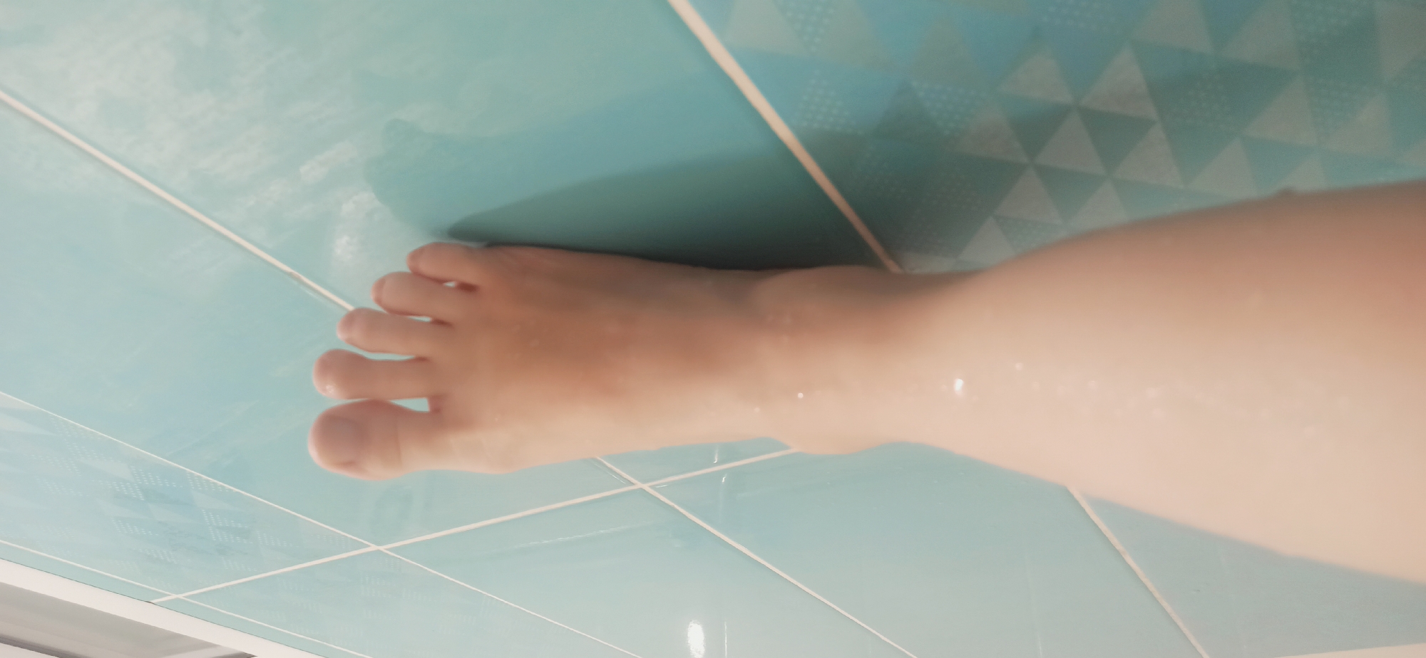 The image shows a close-up view of a person's left foot and lower leg against a background of light blue tiles. The tiles are arranged in a grid pattern, with white grout lines. The foot and leg appear wet, with small droplets of water visible on the skin. The lighting suggests the photo was taken indoors, possibly in a bathroom. The image does not contain any elements that would suggest a specific location within the city of Asbest, Russia.