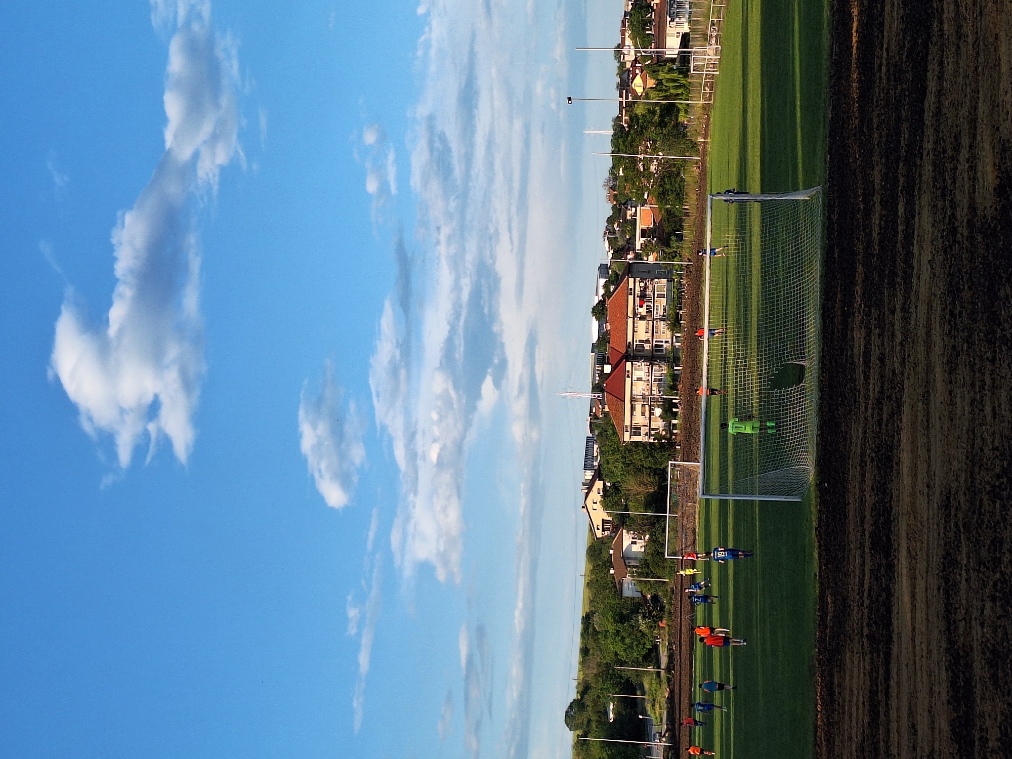 An outdoor football pitch in Ezerovo, Bulgaria, is observed under a bright blue sky with scattered white cumulus clouds, suggesting a late afternoon or early evening setting due to the soft, warm light. In the immediate foreground, a strip of dark, disturbed earth with visible indentations occupies the lower portion of the frame. A white football goal with a net stands prominently on the right side of the midground. On the green grass pitch, multiple individuals are engaged in a football game or practice. One player in a bright green uniform is positioned within the goal area, facing away from the camera. Other players are dressed in blue and orange/red jerseys, with one blue-jerseyed player featuring the number "19" on his back. A second white goal is visible at the far end of the pitch. Beyond the playing field, a dense line of green trees and bushes separates the pitch from a residential area. Several multi-story houses with light-colored facades and red-tiled roofs are scattered across the background. Further back, a white cellular or communication tower rises above the distant structures. Utility poles and power lines are visible, particularly on the left side of the background, alongside what appears to be a road.