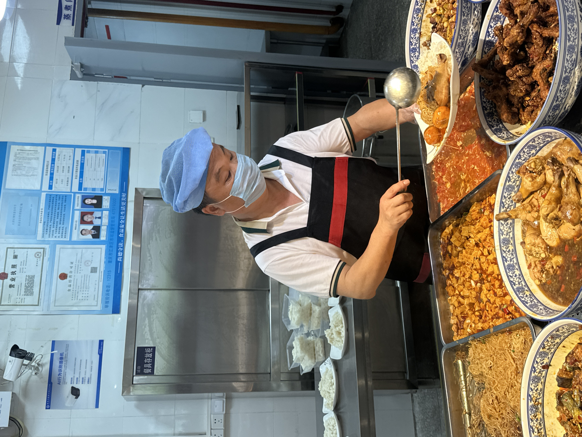 Here is a matter-of-fact description of the scene:
The image shows an indoor food service environment, likely a cafeteria or canteen, in the city of Jiangjin, China.
A person, wearing a white shirt with dark trim, a black apron with a red stripe, a light blue hairnet, and a face mask, is using a large metal ladle to serve food from a display containing multiple dishes. The food includes dishes of braised chicken pieces, and fried fish, and other unidentified cuisine. The dishes are presented in white and blue porcelain bowls and rectangular metal trays.
Behind the food server is a stainless steel surface, possibly a countertop. Above it, affixed to a white tiled wall, are various documents, including a business license and a food business permit, with Chinese text. Also visible is a sign mentioning "AI behavior recognition camera" and a sign indicating "utensil storage cabinet" in Chinese. The sign includes the Jiangjin District Market Supervision Administration telephone number.