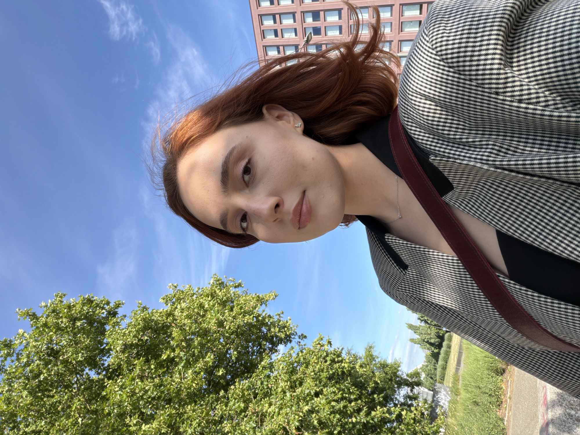 A young Caucasian woman with reddish-brown hair is taking a selfie outdoors on a sunny day with a blue sky and scattered clouds. She is wearing a black top, a black and white checkered blazer, and a burgundy strap from a bag across her chest. A silver necklace with a small pendant is visible. She has on a small, silver stud earring in her right ear. In the background to the left, there is a large, leafy green tree. To the right, a modern pink building with many windows is visible. In the far background, there are more trees and some grass. The scene appears to be in an urban or suburban environment, possibly in Strasbourg, France, given the architectural style of the building.