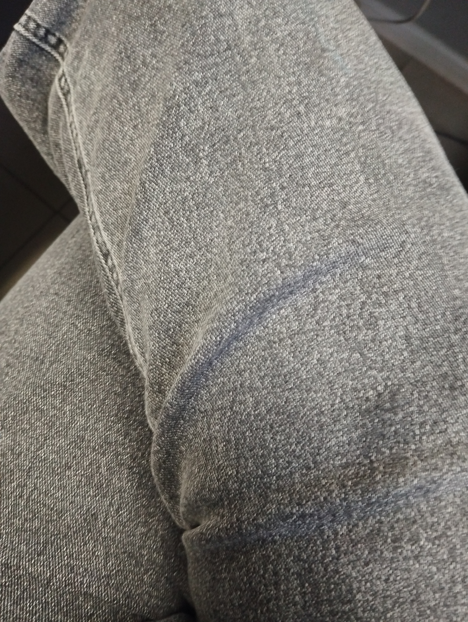 A close-up, top-down shot shows a pair of dark grey denim jeans. The fabric is textured with the characteristic weave of denim. A subtle crease runs diagonally across the right side of the frame, suggesting the material has been folded or worn. The stitching along a seam is visible on the left side, running vertically. The lighting creates some highlights and shadows, emphasizing the texture of the fabric. The background is dark and out of focus. This image is a detail of clothing, not a scene that can be located in a specific city or environment.