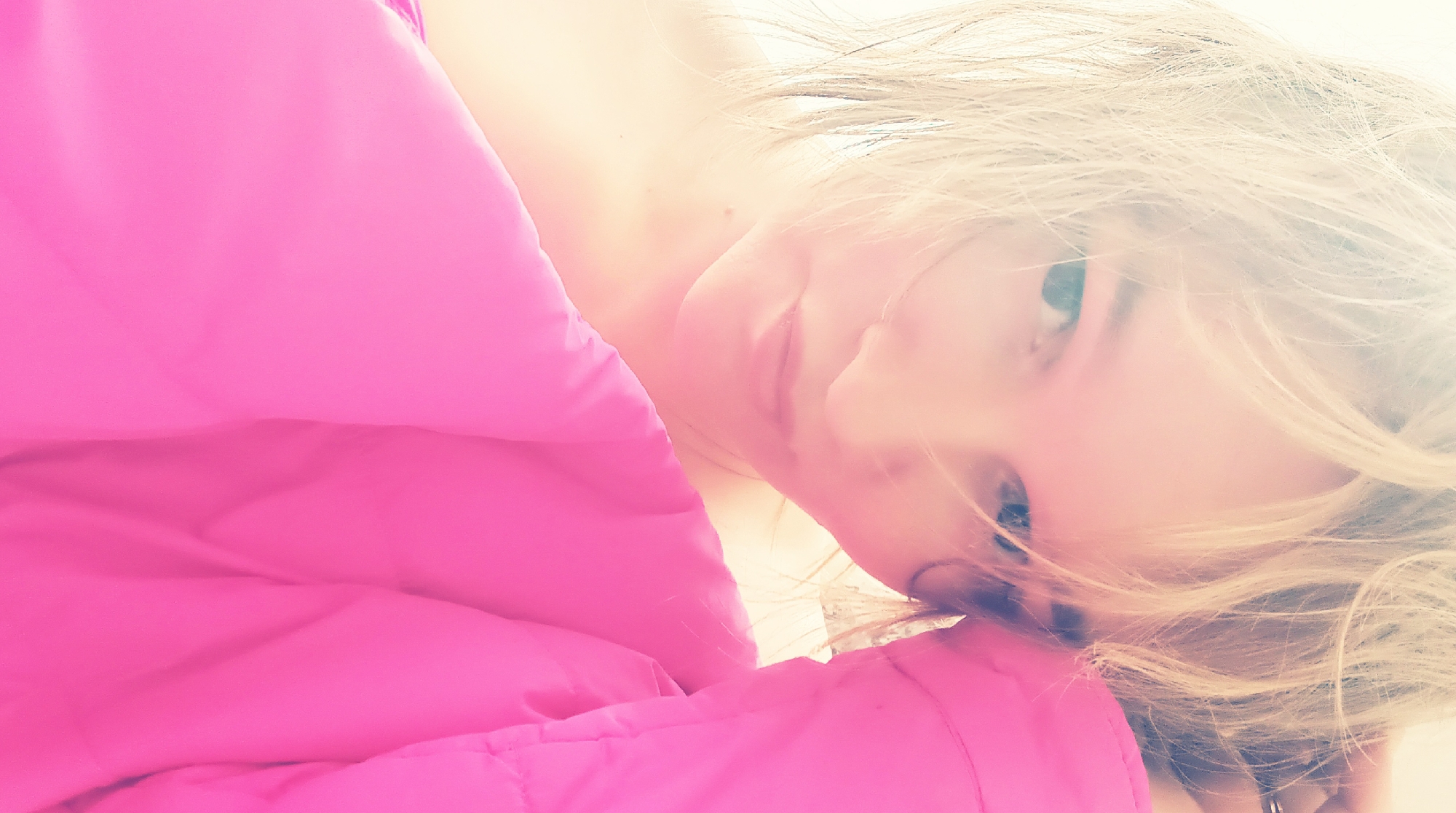 A brightly lit, close-up portrait depicts an adult female from the upper torso up. She has blonde, disheveled hair that partially covers her forehead and temples. Her dark eyes are directed towards the viewer, and her mouth is closed with a neutral expression. A bright pink, possibly padded, garment is draped over her left shoulder and across the lower right portion of the frame. A light-colored ring is visible on her left ring finger, partially obscured by hair. Her left hand is raised, with fingers near her head. The background is an indistinct, overexposed white area, indicating an indoor setting with a strong, diffuse light source. The specific location within the city of Ольховка, Russia, cannot be determined from the visual information provided.