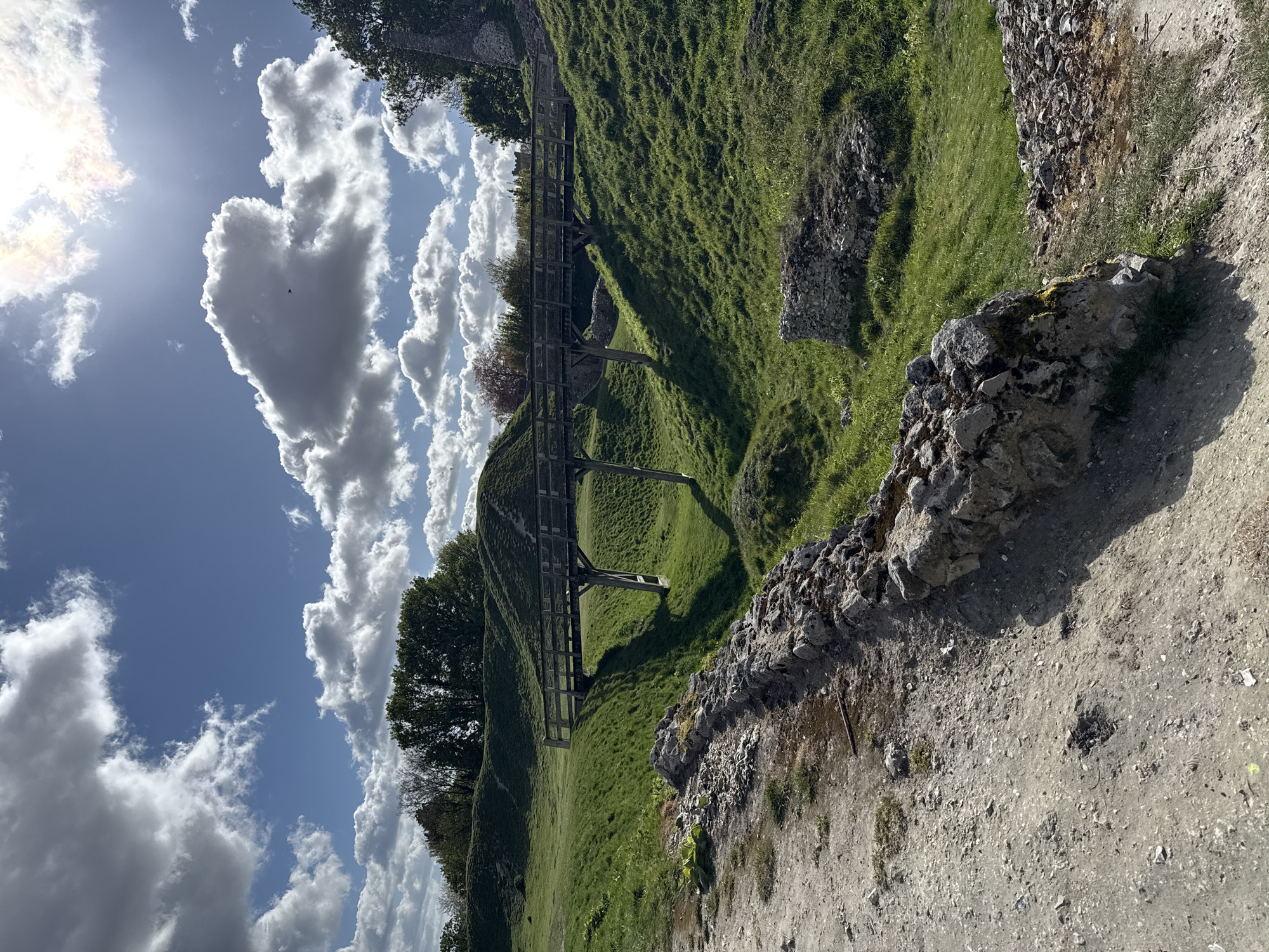 The image captures a vibrant outdoor landscape featuring undulating green terrain, likely earthworks or mounds, under a bright, partly cloudy sky. The scene is set in what appears to be a historical or natural park in King's Lynn and West Norfolk, United Kingdom.
In the mid-ground, a wooden footbridge or walkway with railings spans across a dip in the grassy mounds, connecting two higher points. The slopes of the mounds are covered in lush green grass, illuminated by the sun, creating varied shades and shadows. Scattered trees are visible in the distance and nestled among the mounds, adding to the natural environment.
In the immediate foreground and extending along the right side of the image, there is a rough stone wall or the remnants of a structure, suggesting ancient ruins or a boundary. Adjacent to this wall is a dirt or gravel path, hinting at pedestrian access.
No people are visible, but a small, dark bird can be seen soaring against a large cloud in the sky.
The time of day is bright, likely late morning or early afternoon, with the sun shining intensely from the upper left, casting strong shadows from the wooden bridge and terrain features. The weather is clear and fair, characterized by a blue sky dotted with large, puffy white cumulus clouds, creating a dynamic backdrop. The overall impression is one of tranquility and natural beauty, characteristic of a historical or nature preserve. There is no visible text in the image.