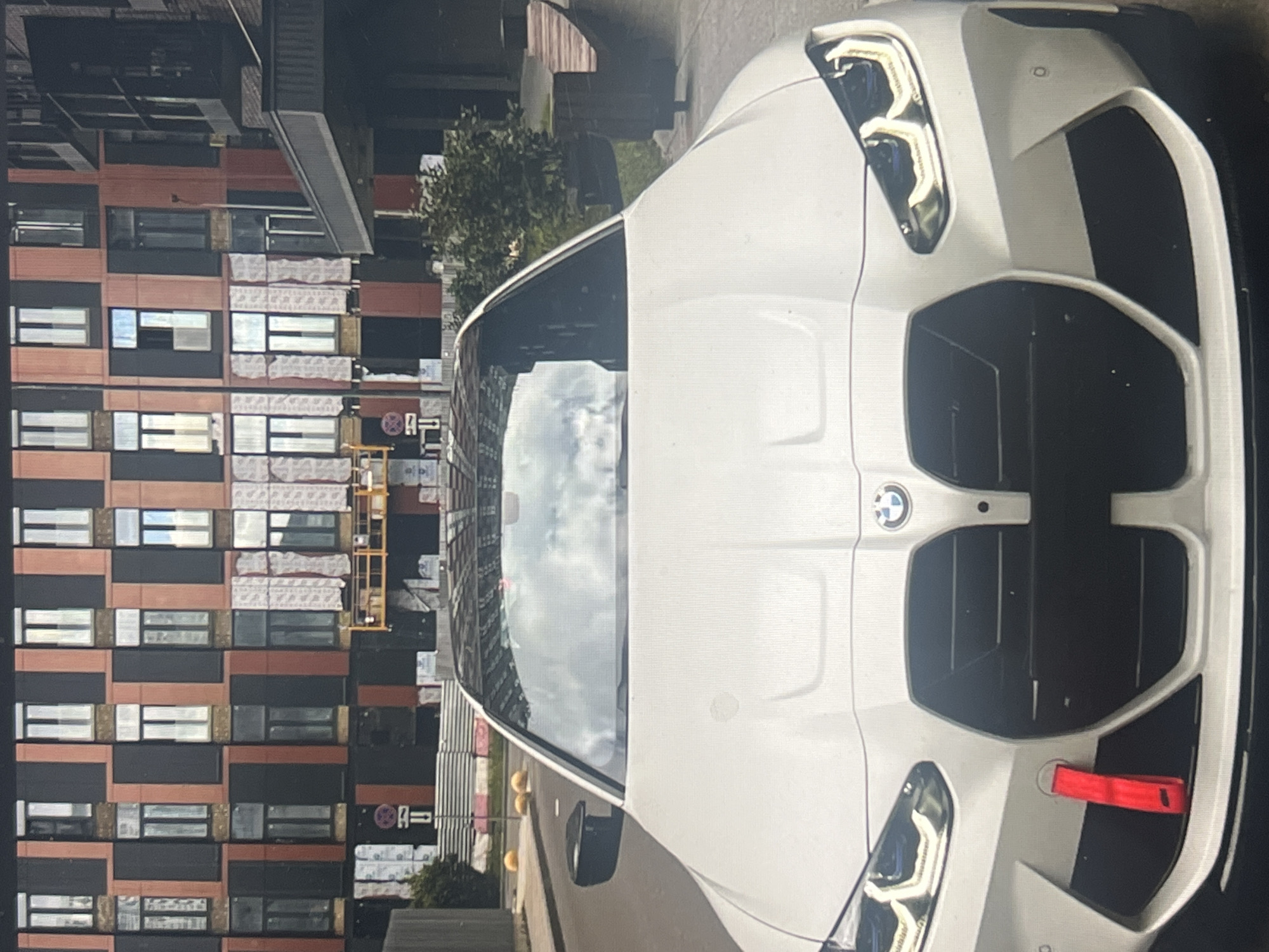 The image shows the front of a white BMW car parked on a street in front of a modern building. The building has a facade with a repeating pattern of reddish-brown and black vertical elements, interspersed with windows. Some sections of the building appear to be under construction, with white protective sheeting visible. Yellow scaffolding is present on the side of the building. In the background, there are trees and a sidewalk. The car has a black grille, distinctive headlights, and a red tow strap attached to the front bumper. The sky is cloudy. The image does not provide any specific information that would allow for the determination of its location in Vanadzor, Armenia.