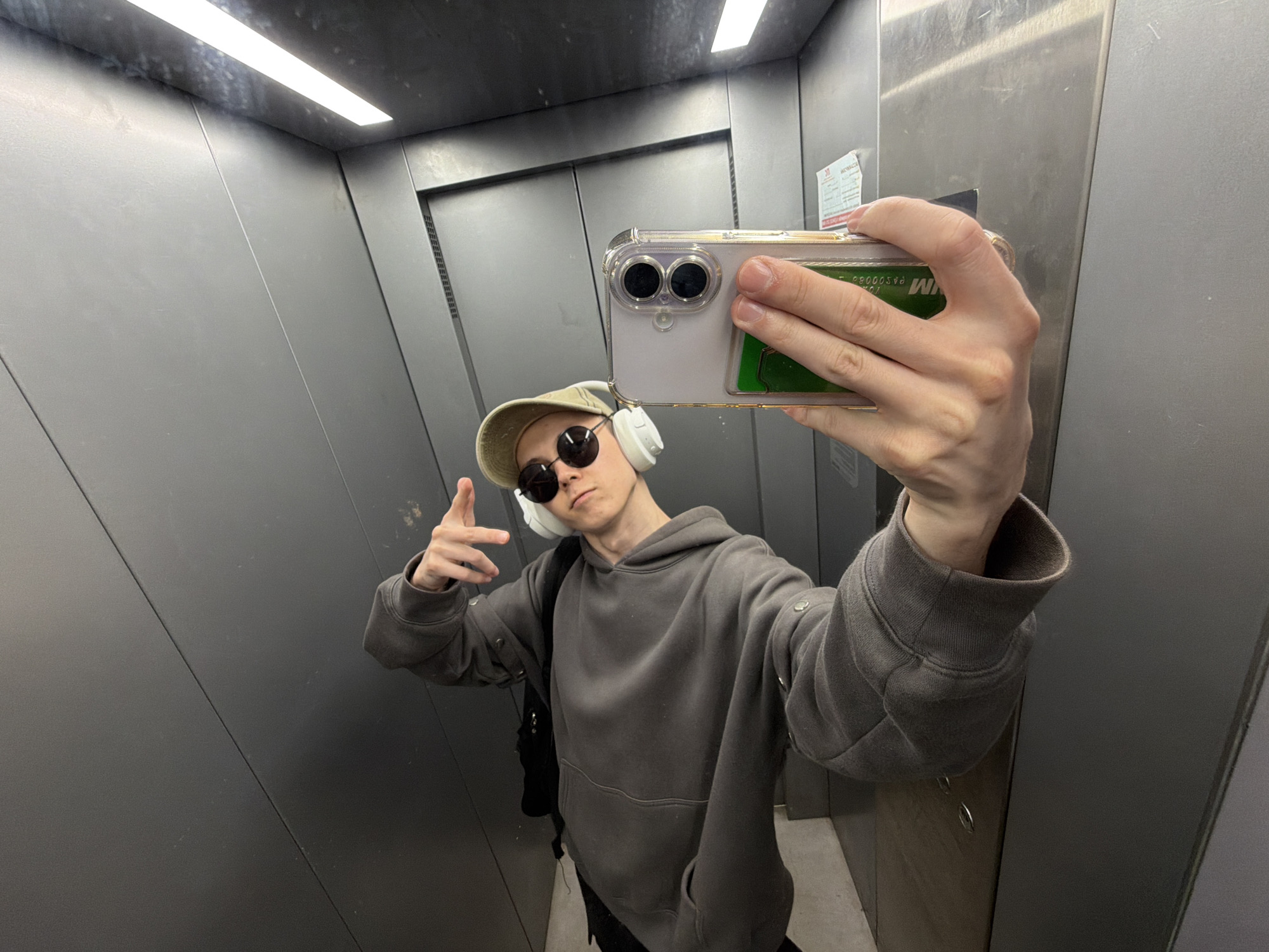 A person is taking a selfie in an elevator. The person is wearing a grey hoodie, a light-colored baseball cap, round sunglasses, and white headphones. They are holding a smartphone in their right hand, with the camera facing the mirror. The elevator walls are made of brushed metal, and a fluorescent light illuminates the space from above. The person is making a peace sign with their left hand. The phone case has a green card sticking out of it. The overall mood is casual and relaxed. There is some text visible on a sticker on the elevator wall behind the person. The time of day and weather are not discernible from the image.