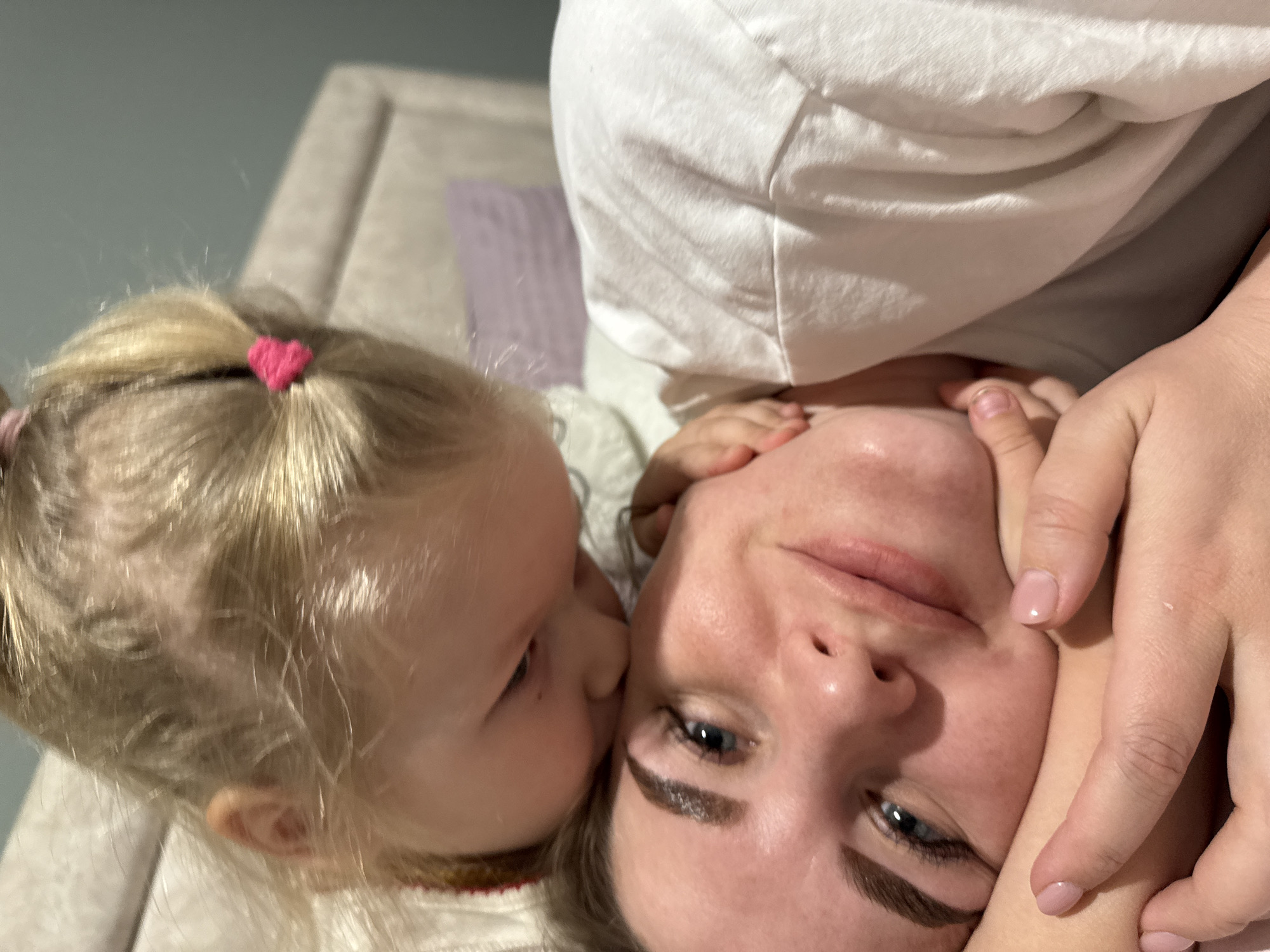 A close-up shot shows a woman lying on what appears to be a bed, with a young child resting their head against her cheek. The child has blonde hair secured with a pink heart-shaped hair tie. The woman has light skin, blue eyes, and manicured nails. Her hand is positioned near her face. The setting seems to be indoors, based on the presence of a headboard visible in the background. The location in the city of Gornyi (Dvurechenskoe S/P), Russia, is not possible to determine from the provided image.