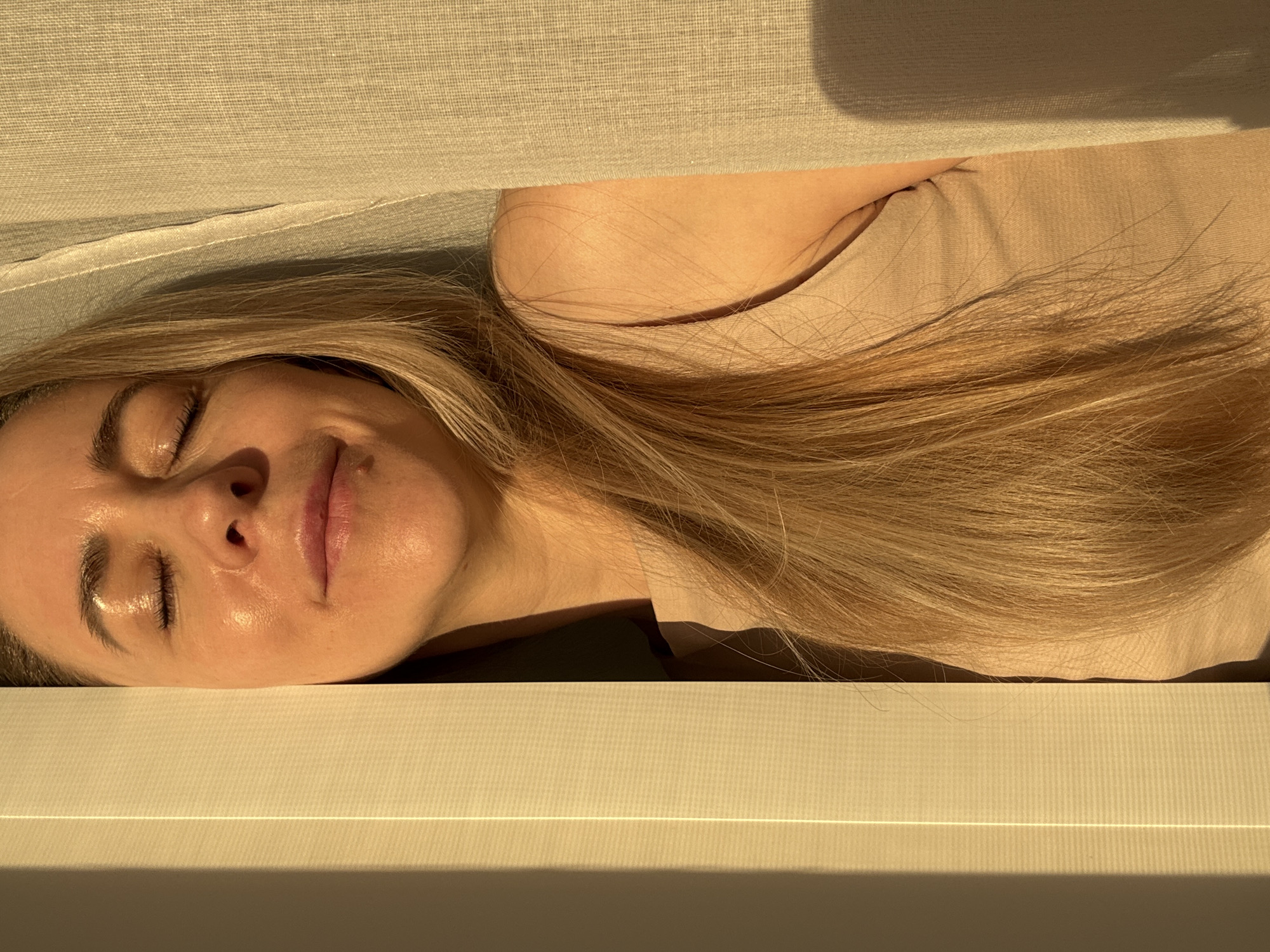A woman with long blonde hair, light skin, and closed eyes lies on a cream-colored surface. She is facing upwards, with a faint smile on her lips. A fabric, possibly a curtain or sheet, is draped above her. The image has a warm, golden light, suggesting it's near sunrise or sunset. The location is not identifiable from the image.