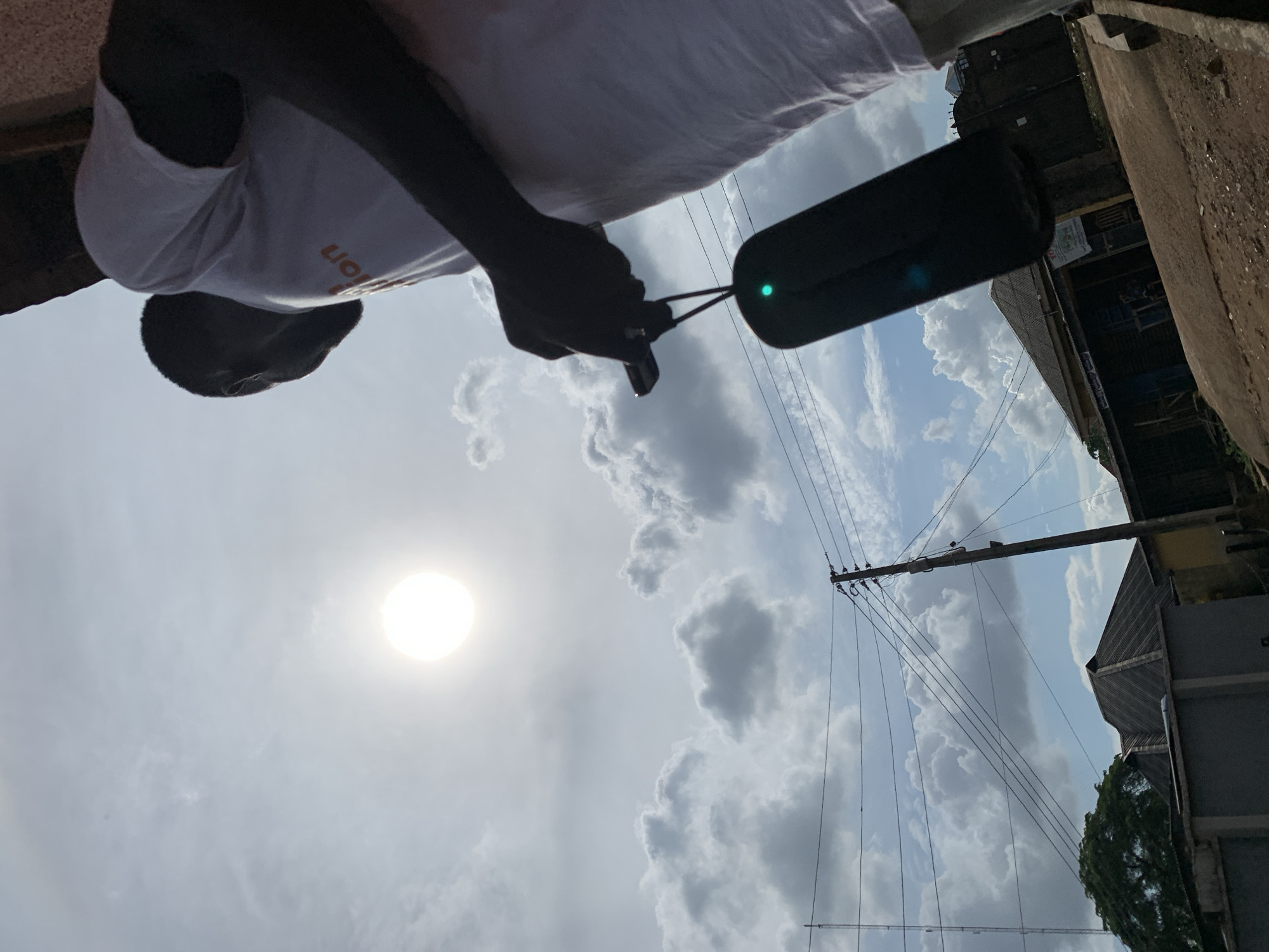 The image is taken from a low angle, looking upwards. A person, possibly the photographer, is visible wearing a white shirt and holding a black object connected by a thin wire to what appears to be a pair of earphones or a similar device. The person's face is dark.
In the background, there is a bright sun obscured by clouds. Electrical poles and wires stretch across the sky. Buildings are visible along the right edge of the frame, partially obscured, along with greenery.
Based on the location provided, the scene appears to be a street view in Ikorodu, Nigeria, characterized by power lines, buildings, and a partially cloudy sky.