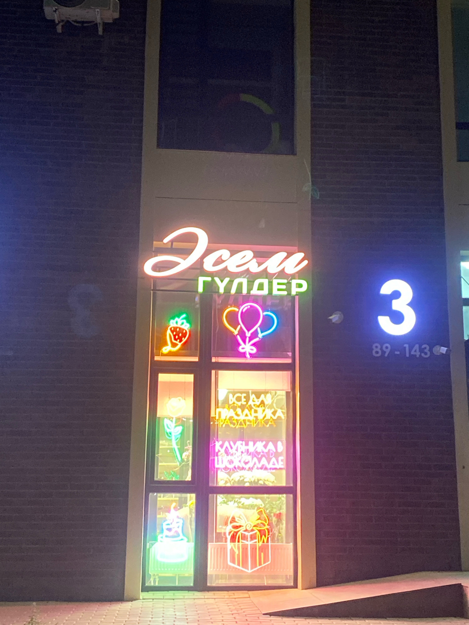 An exterior nighttime photograph captures a neon-lit storefront at number 3 on an unspecified street in Astana, Kazakhstan. The facade of the building is constructed of dark brick. Above a storefront window, a neon sign reads "Всем ГУЛДЕР" in stylized script and block letters, with the first word in pink and the second in green. Within the storefront window, illuminated by neon lights, are various decorative elements. In the upper left section, a neon strawberry and two neon balloons are visible. Below these, a neon rose and another neon item that appears to be a cake are displayed. The right side of the window features neon text that reads "ВСЕ ДЛЯ ПРАЗДНИКА" (All for the holiday) and "КЛУБНИКА В ШОКОЛАДЕ" (Strawberries in chocolate). A neon gift box is also visible in the lower right section of the window. To the right of the storefront, a large, illuminated blue numeral "3" is mounted on the brick wall, with smaller text below that reads "89-143". A paved walkway with a ramp leading up to the storefront is in the foreground. An air conditioning unit is mounted on the wall above the window.