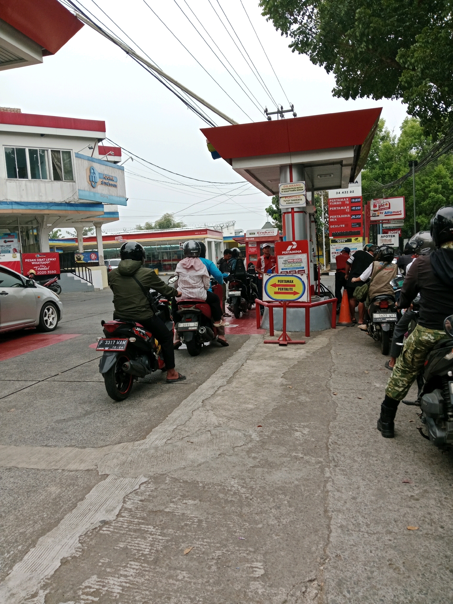 The image shows a queue of motorcycles and cars at a Pertamina gas station in Muka, Indonesia. A silver car is parked on the left, and several motorcycles are lined up to refuel. People in helmets are seen riding the motorcycles, some in casual attire and others in what appears to be a uniform. One person in a green jacket is on a motorcycle, facing away from the camera. Another person in a pink top and helmet is on a motorcycle behind them. The gas station has a red and white canopy over the pumps, with signage indicating fuel prices and types. There are also signs advertising products and services from Alfamart and other businesses. The environment appears to be an outdoor urban setting with trees visible in the background. The sky is overcast.
