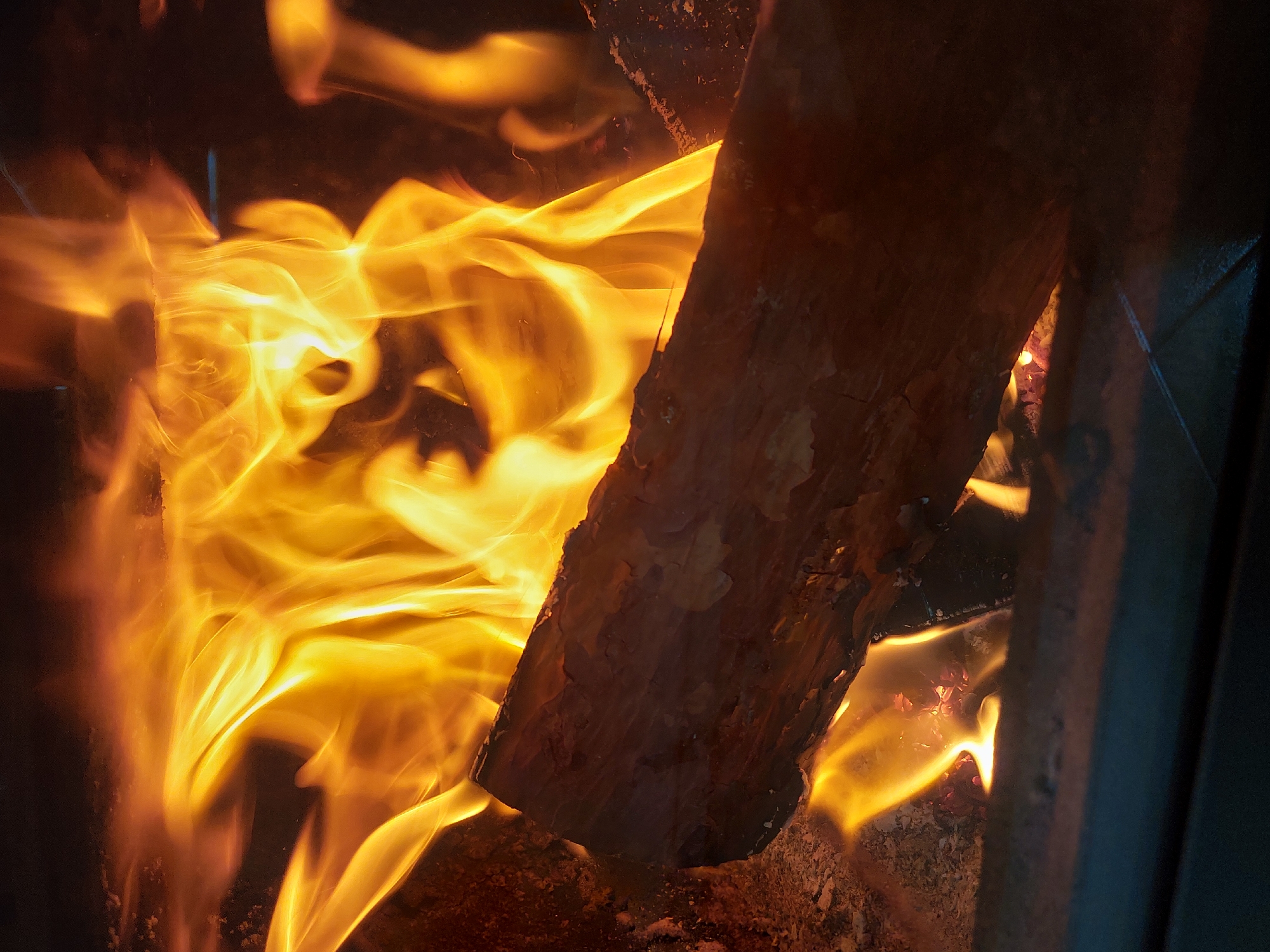 The image shows a close-up view of a burning log in a fireplace. The flames are bright yellow and orange, swirling around the log, which is dark brown with visible bark. The environment appears to be dimly lit, with dark shadows surrounding the fire. There are no people visible in the image. The location of Nedelino, Bulgaria is not evident from the visual information present.