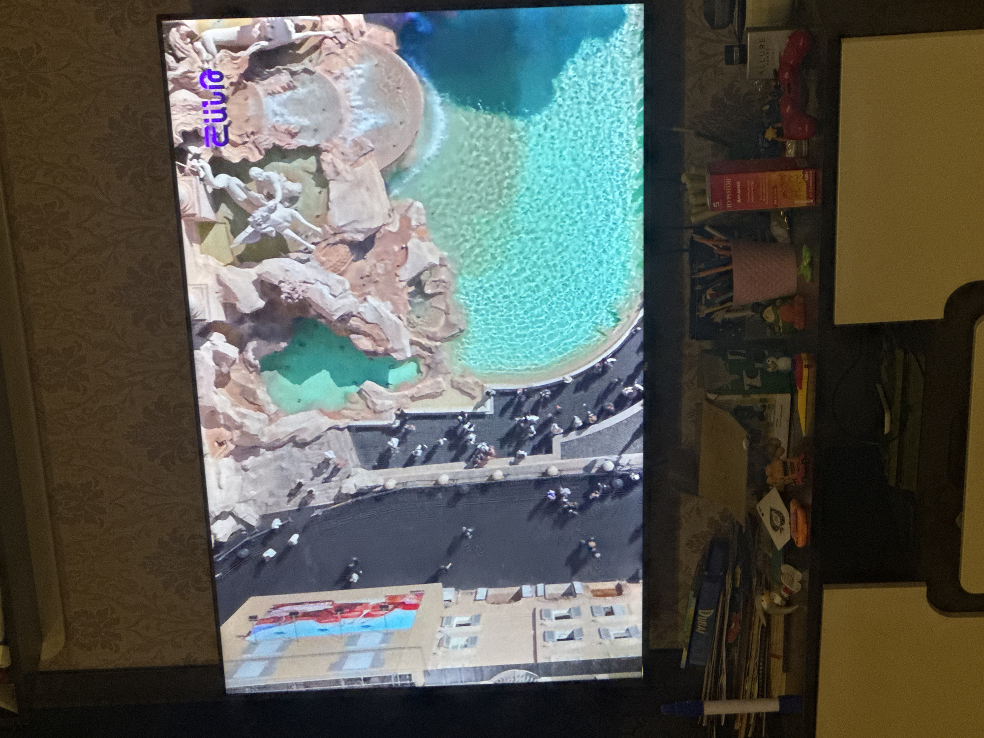 The image shows a television screen displaying an aerial view of the Trevi Fountain in Rome, Italy. The fountain's Baroque sculptures are prominently visible, as is the turquoise water of the basin. Numerous people surround the fountain. The bottom of the screen shows adjacent buildings, including a section with a red and blue awning. Off to the right of the screen are various objects on a shelf, including a red game controller, small boxes, cards, and pens. The wall behind the TV has a textured floral pattern. The location is likely a private residence. The scene does not appear to be in Yerevan, Armenia.