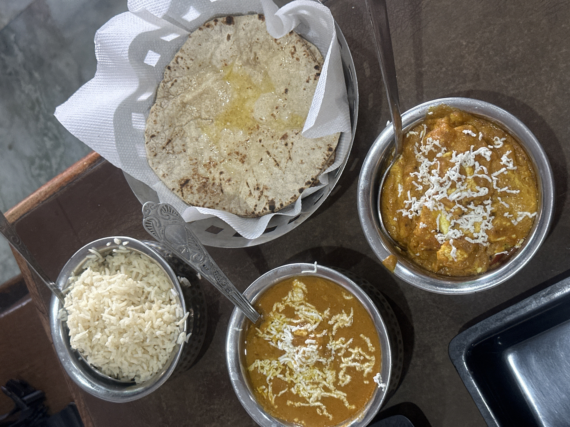 The image shows a meal, likely in Vrindavan, India, consisting of roti in a basket, a bowl of rice, and two bowls of curry topped with shredded paneer or cheese. The food is served in metal bowls, and cutlery is present. The setting appears to be a restaurant or dining area.