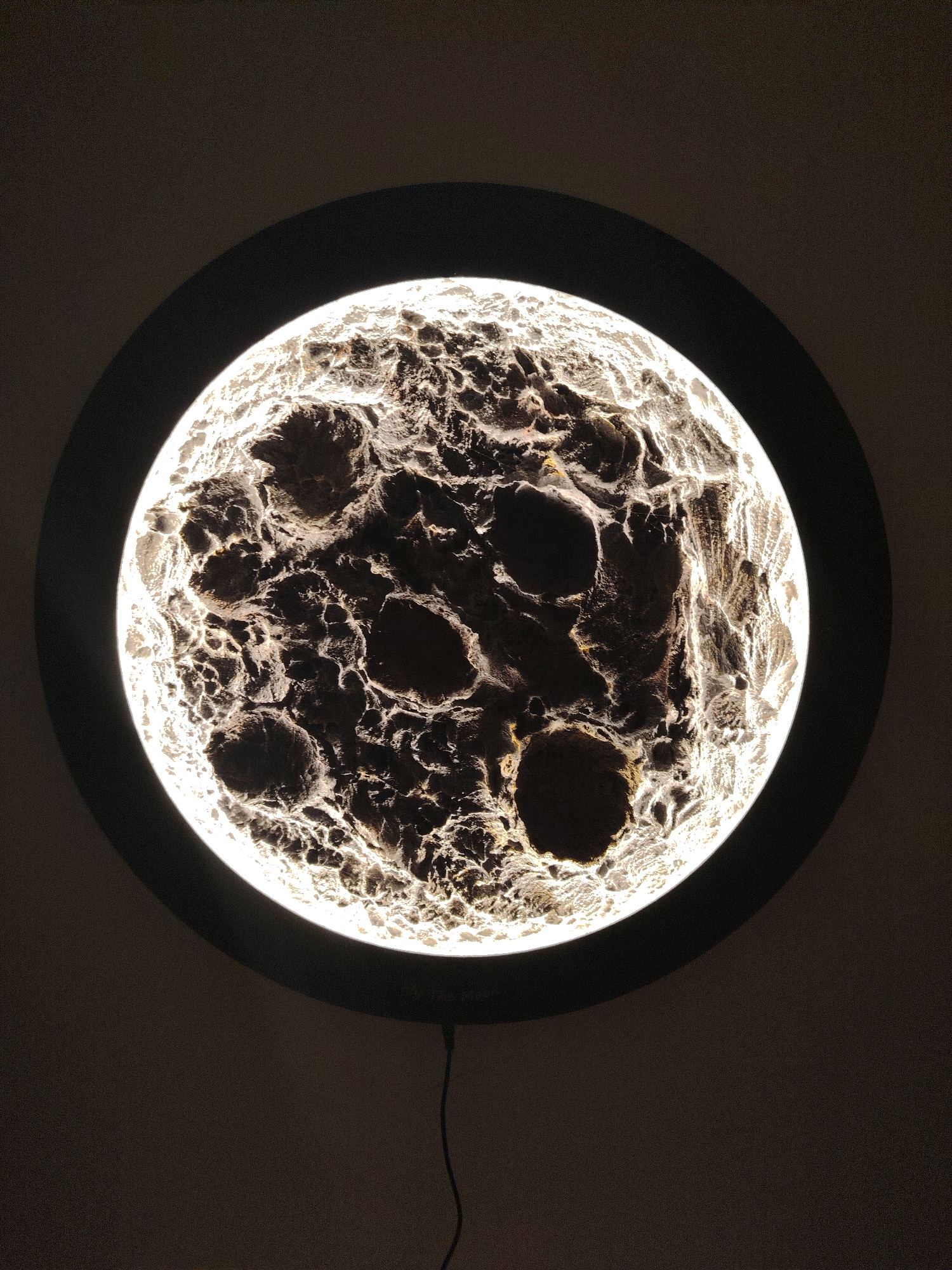 The image presents a circular, internally illuminated display, resembling a heavily cratered celestial body such as the moon. The surface exhibits a rugged, three-dimensional texture with bright, yellowish-white highlights on elevated areas and deep, dark shadows in the depressions and craters. This luminous disk is framed by a thin, matte black circular bezel. The entire object is positioned against a uniform, dark grey background, likely a wall. A thin black electrical cord extends downwards from the bottom center of the black frame, disappearing into the darker lower portion of the background. Faint white lettering, difficult to discern, is visible on the black frame above the cord's origin point. No people, discernible actions, or environmental details that would indicate a specific geographic location in Huntingdonshire, United Kingdom, are present.
