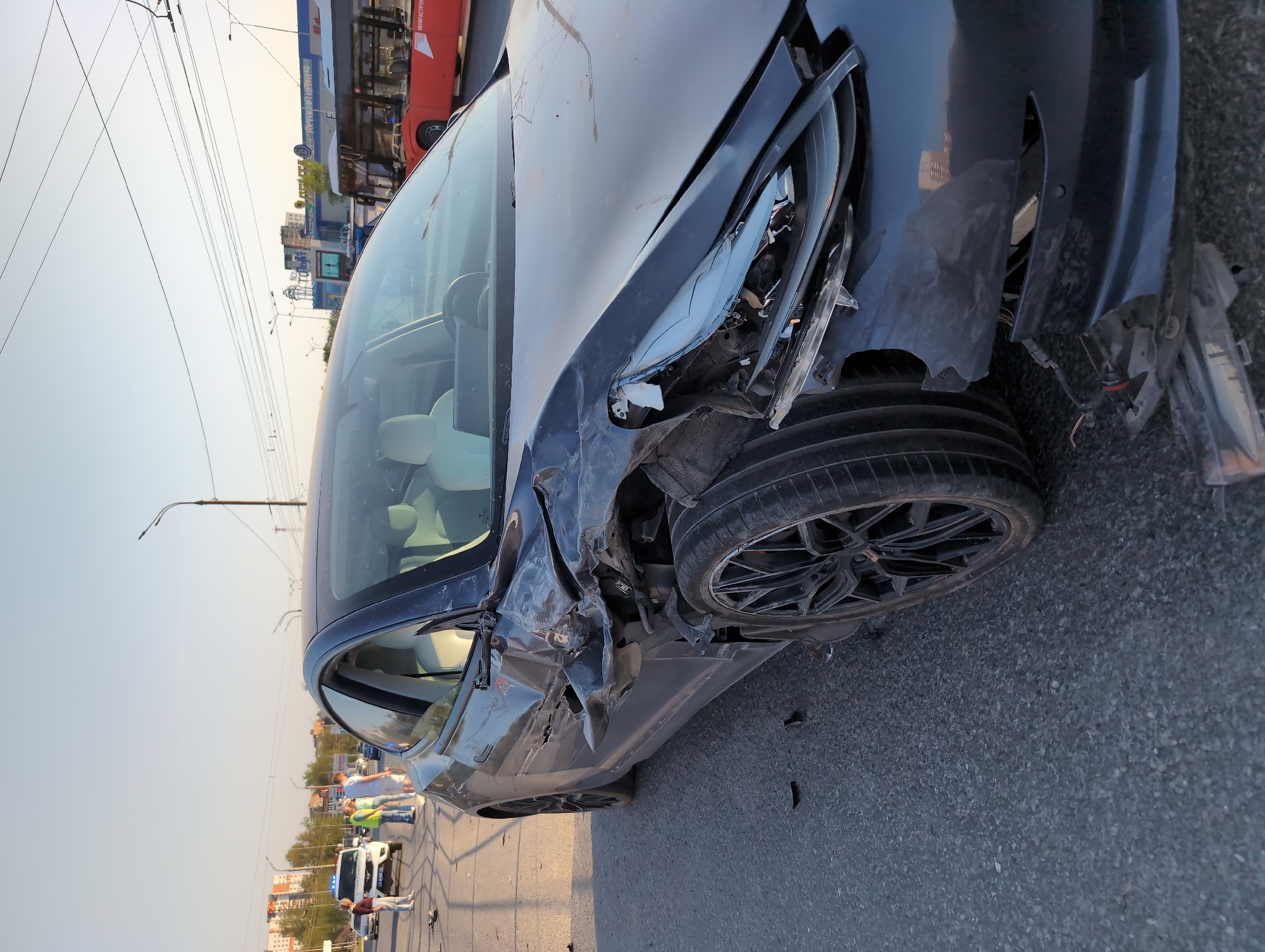 The image depicts a damaged gray car on a paved road, likely a street, in what appears to be an urban or suburban environment. The front end of the car is severely damaged, with broken headlights, a crumpled hood, and a missing front bumper. The front passenger-side tire is visible and appears intact. In the background, there are people standing near a police car with its lights on, suggesting an accident scene. Other vehicles, including a red bus or truck, are also visible in the background. There are overhead power lines. The sky is overcast.