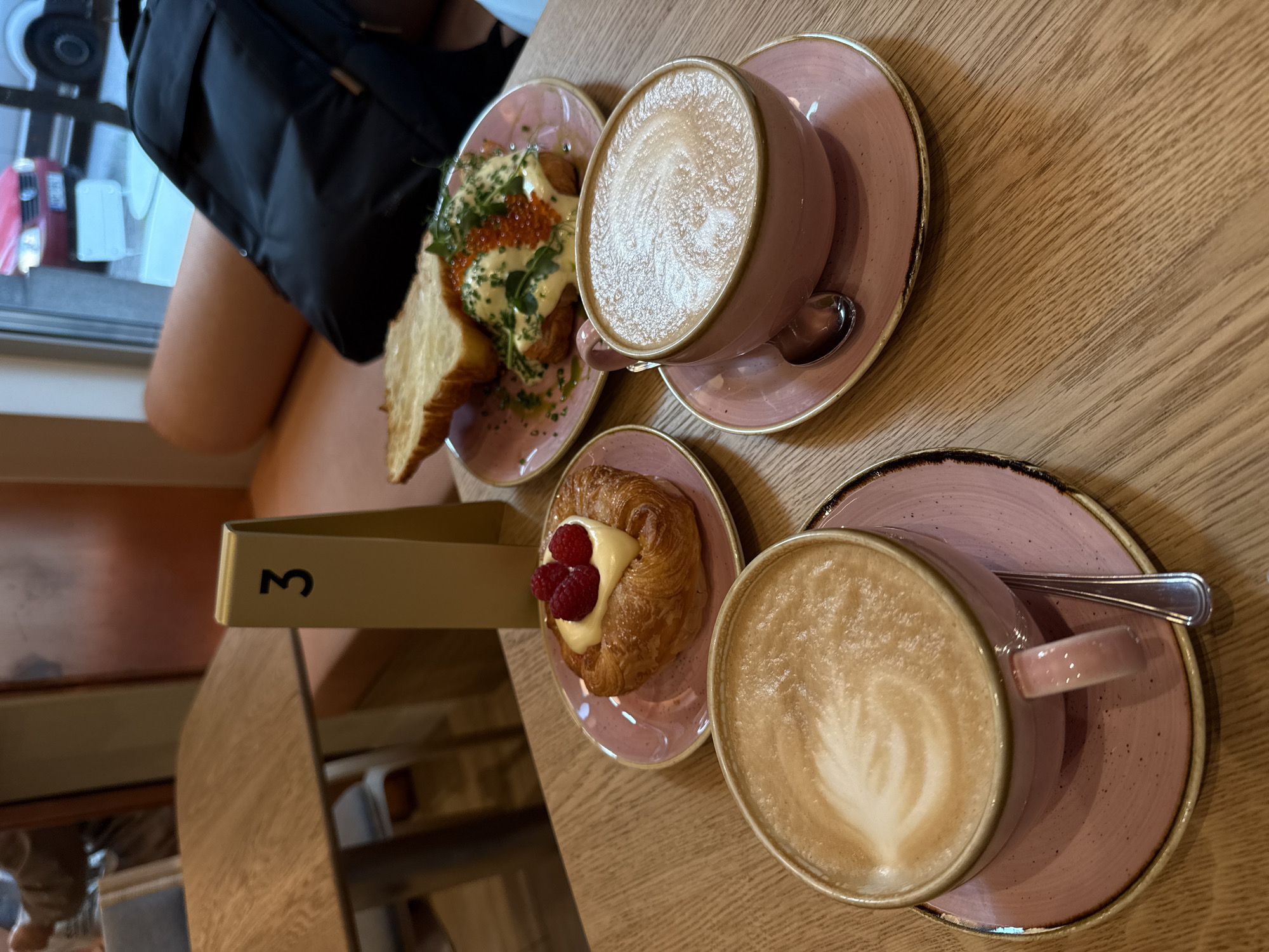 The image shows two pink cups of coffee with latte art on a wooden table at a cafe. To the right of the coffee cups, on a pink plate, are two pieces of toast topped with what appears to be poached eggs, hollandaise sauce, chives, and red caviar. To the left of the coffee cups, on another pink plate, is a pastry with a dollop of cream and three raspberries on top. A gold table number '3' stands behind the pastry. In the background, a black backpack is visible, along with a window showing a street scene with vehicles. The ambiance suggests an indoor cafe setting in Riga, Latvia.